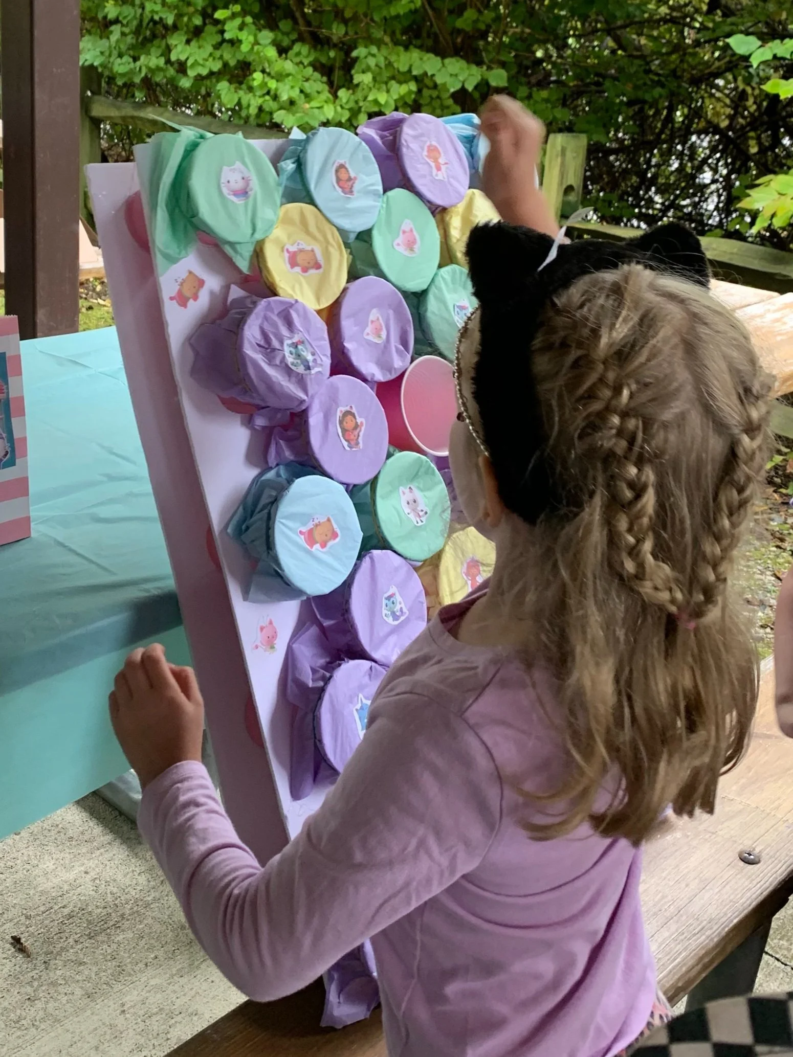 A young girl with braided hair and a black cat on her shoulder playing a ring toss game with colorful cups decorated with cartoon characters.
