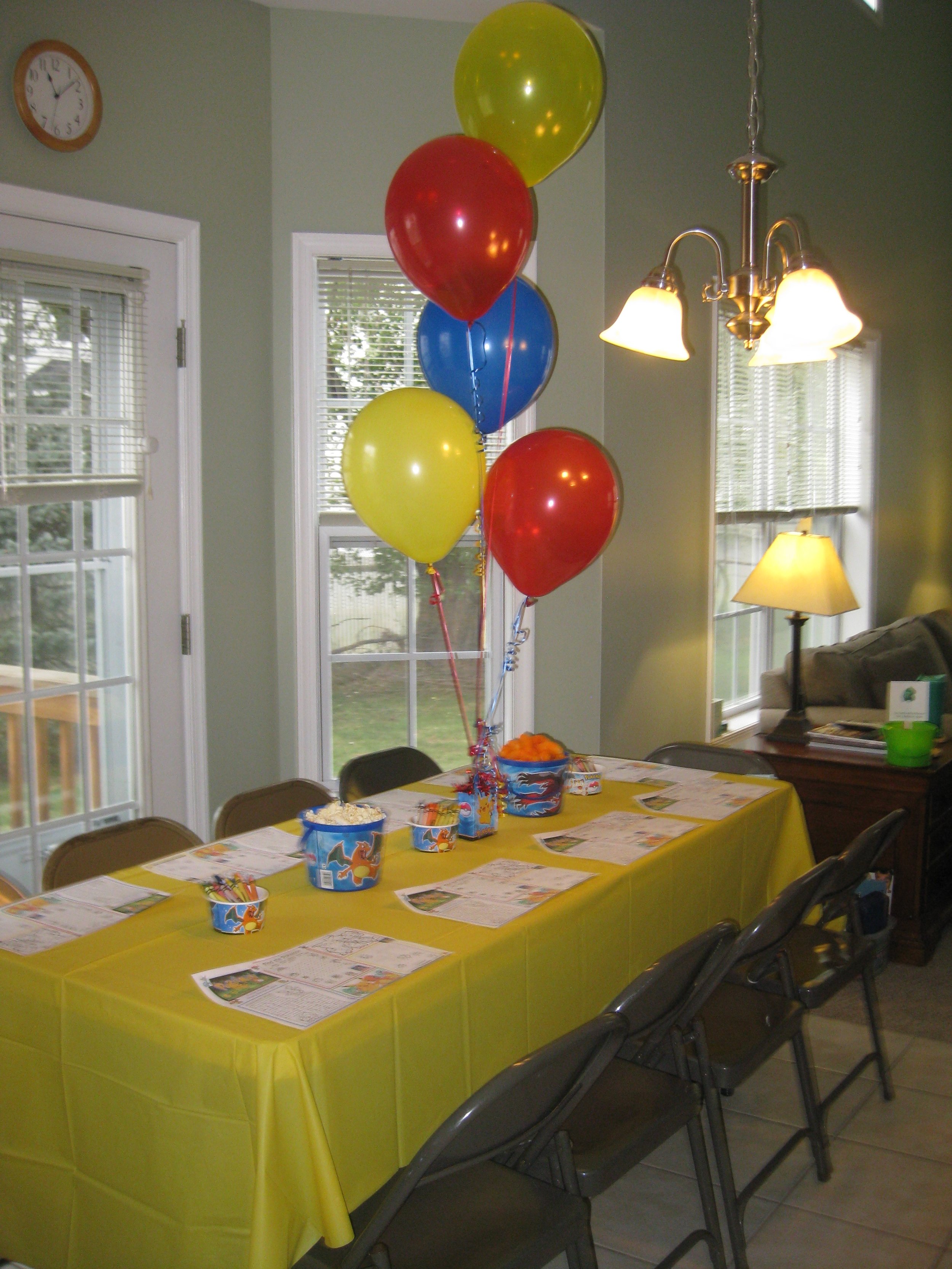 Party table decorated with a yellow tablecloth, colorful balloons, popcorn, and party favors in a dining room with large windows, a ceiling light, and a standing lamp.