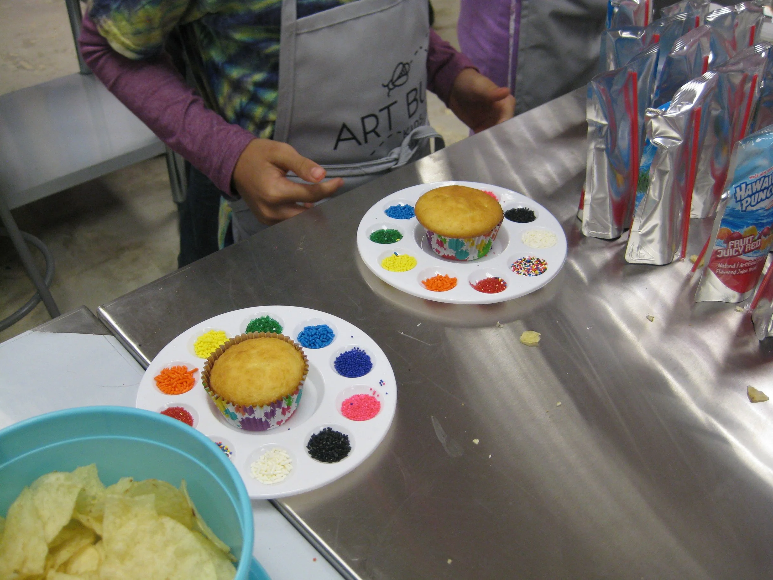 Children decorating cupcakes with colorful sprinkles and frosting, with packs of fruit juice on the table.