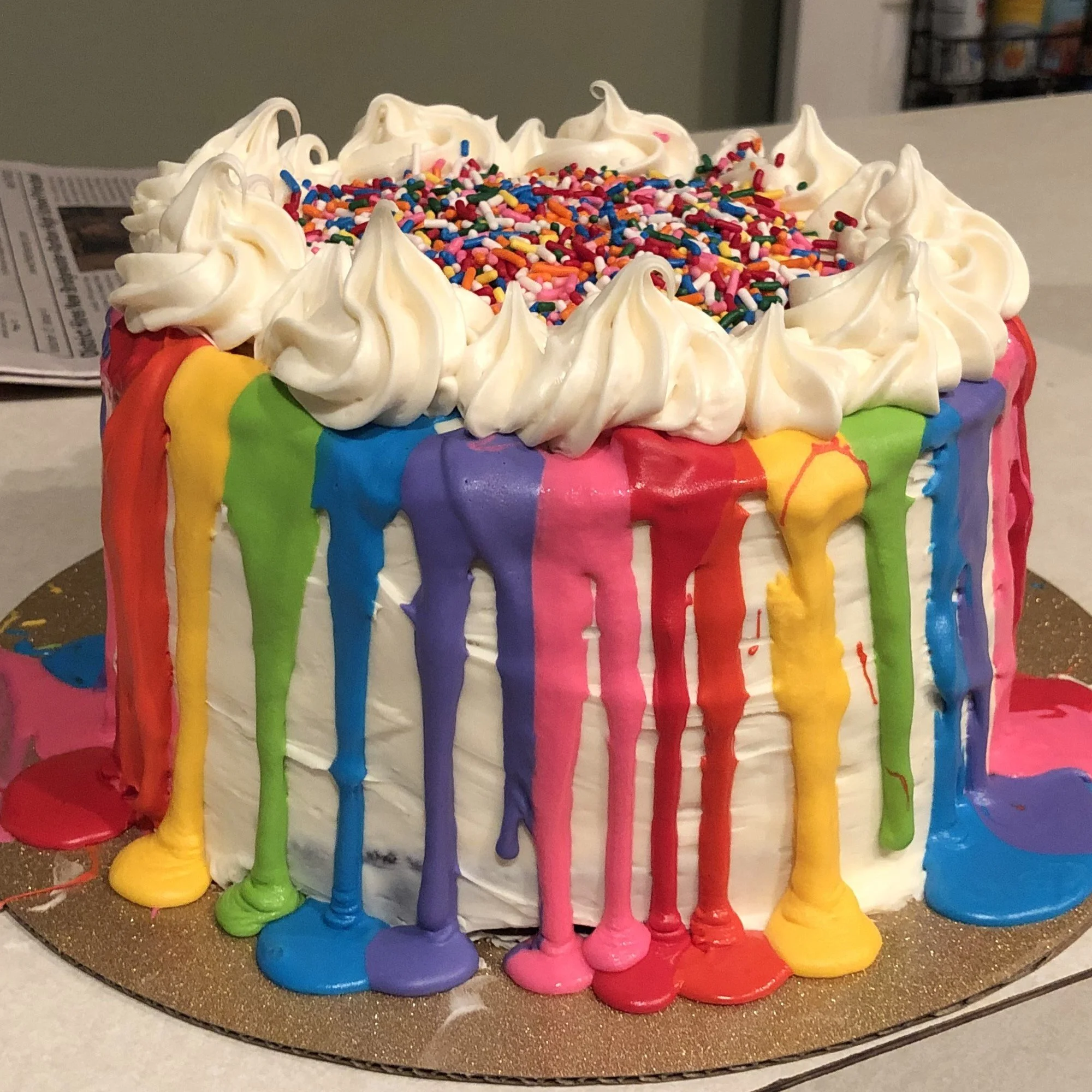 A birthday cake decorated with rainbow-colored icing, rainbow sprinkles on top, and white whipped cream around the edges.