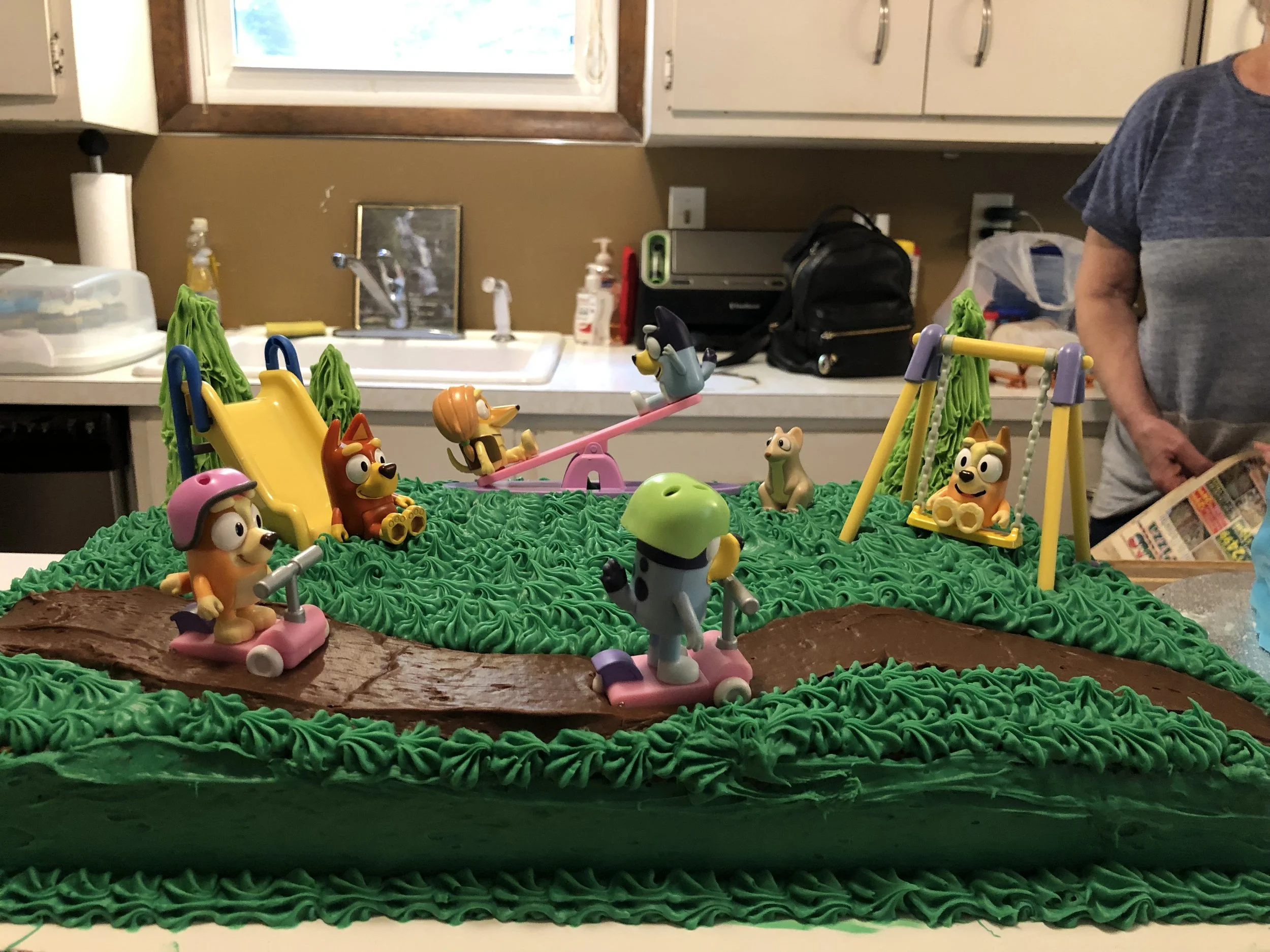 Animal-themed birthday cake decorated with green piped icing, small figurines of dogs, a slide, swing, and seesaw, all resembling characters from a cartoon or show, placed on top of the cake in a kitchen setting.