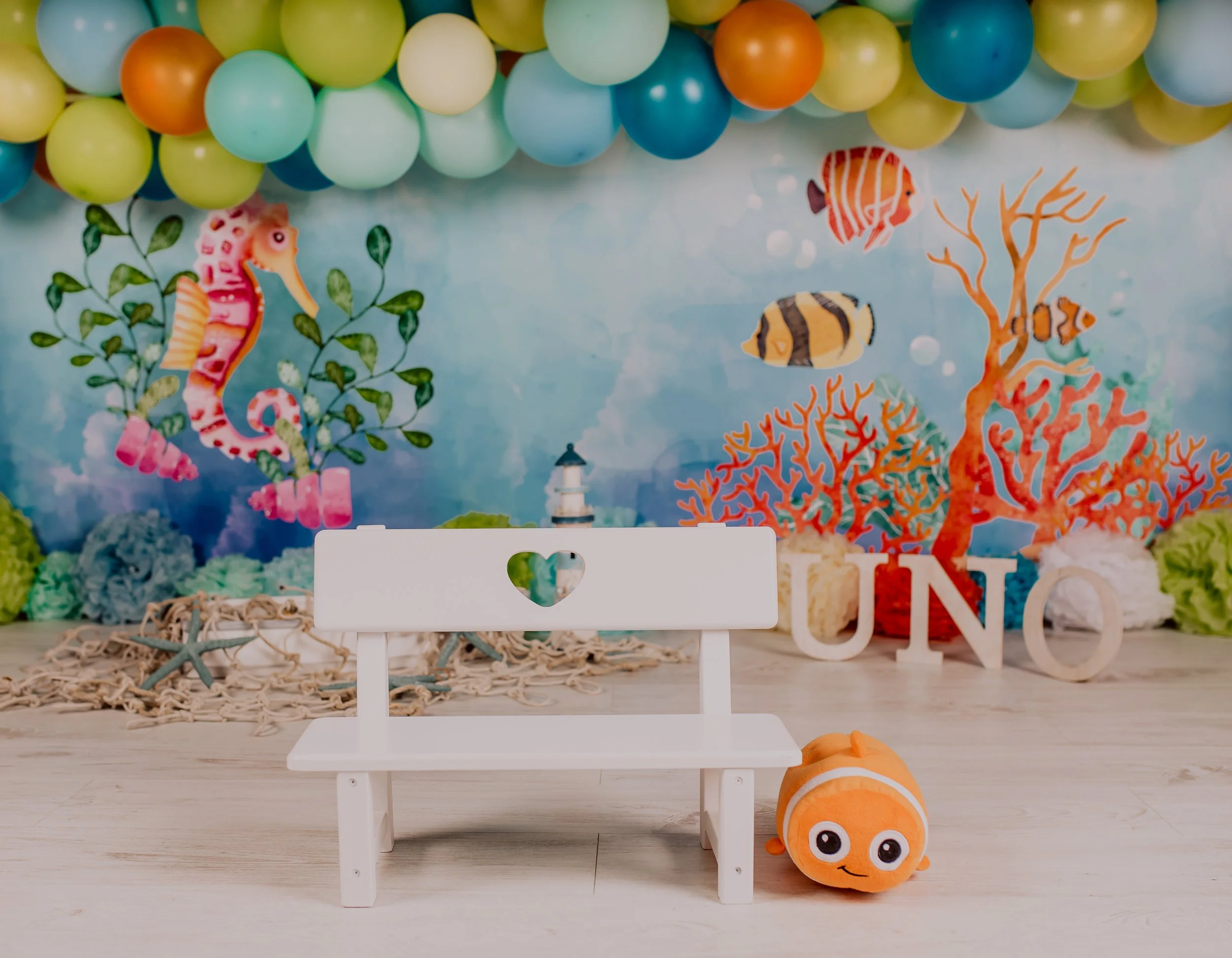 Underwater themed decorations with a backdrop of colorful fish, coral, and a seahorse, a white wooden bench with a heart cutout on the backrest, a plush clownfish toy, and the word "UNO" spelled out in large white letters on the floor.