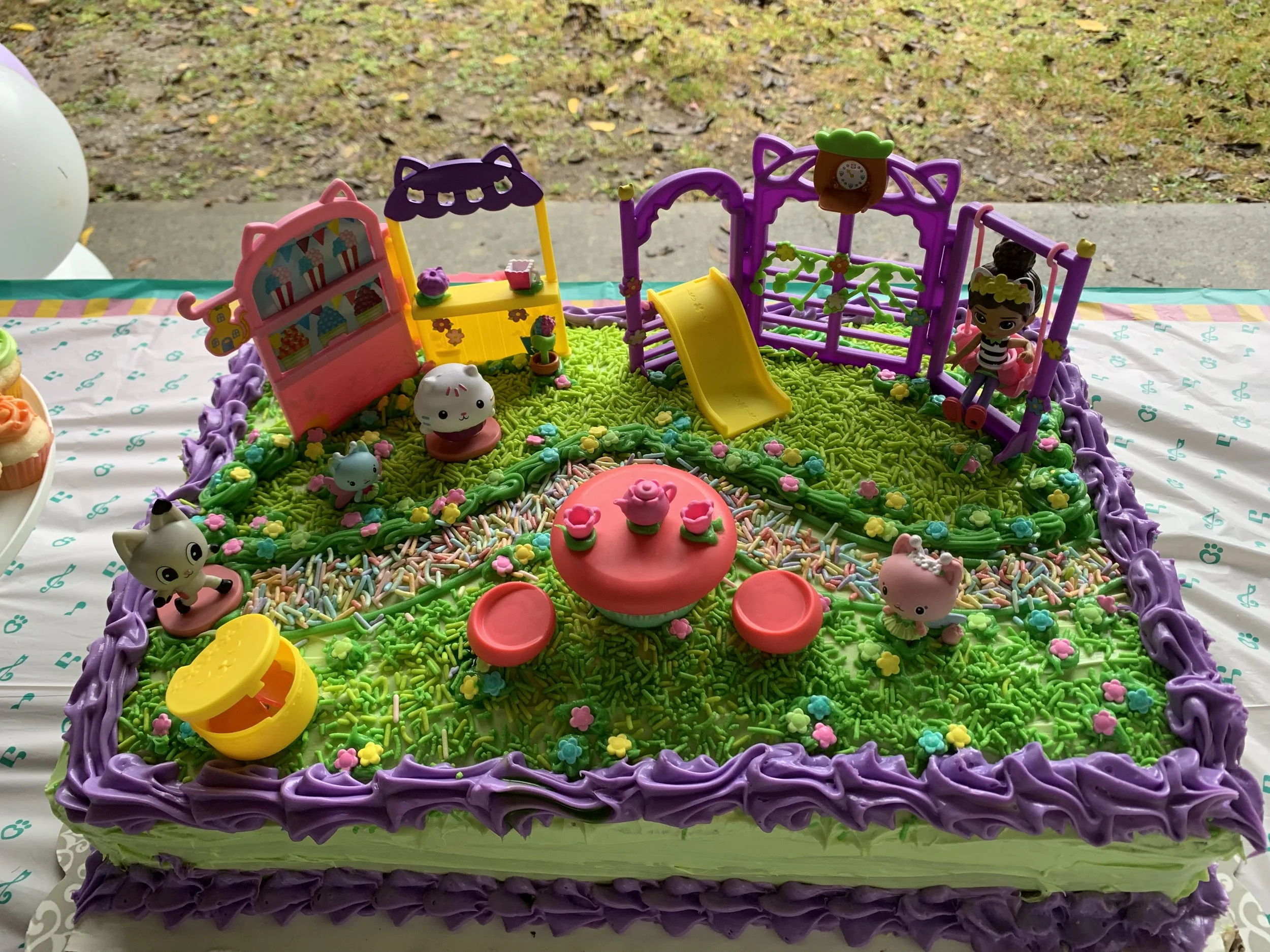 Colorful cake decorated with small toy animals and a miniature playground, including a slide, swing, and animal figures, on a table with a white tablecloth with teal musical note patterns.