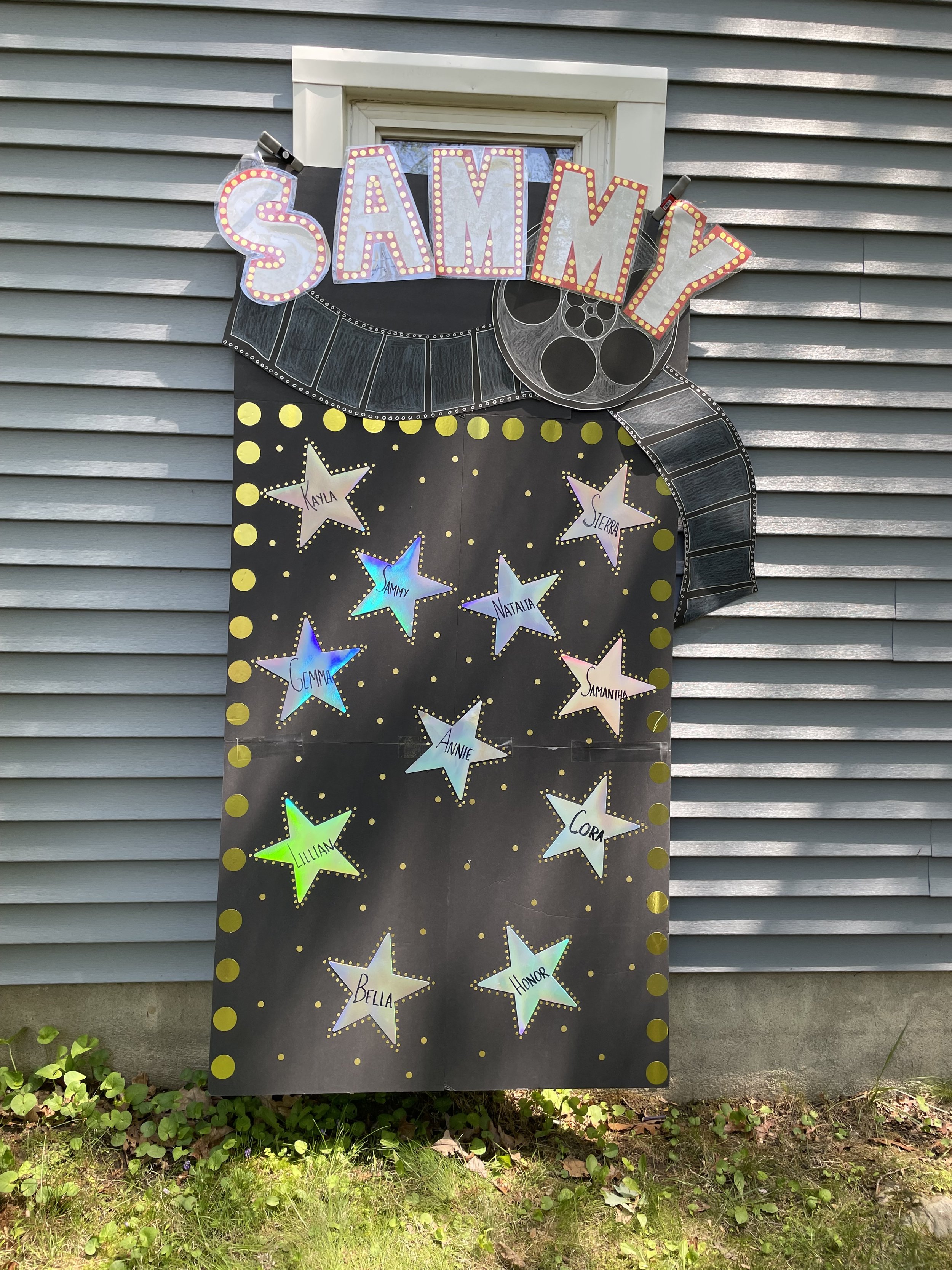Decorative bulletin board with a crescent shaped film reel design at the top, the word 'SAMMY' in large lights above it, and colorful star-shaped cutouts with names written on them pinned to the board. The background is a black paper with gold dots, mounted on a house with gray siding.