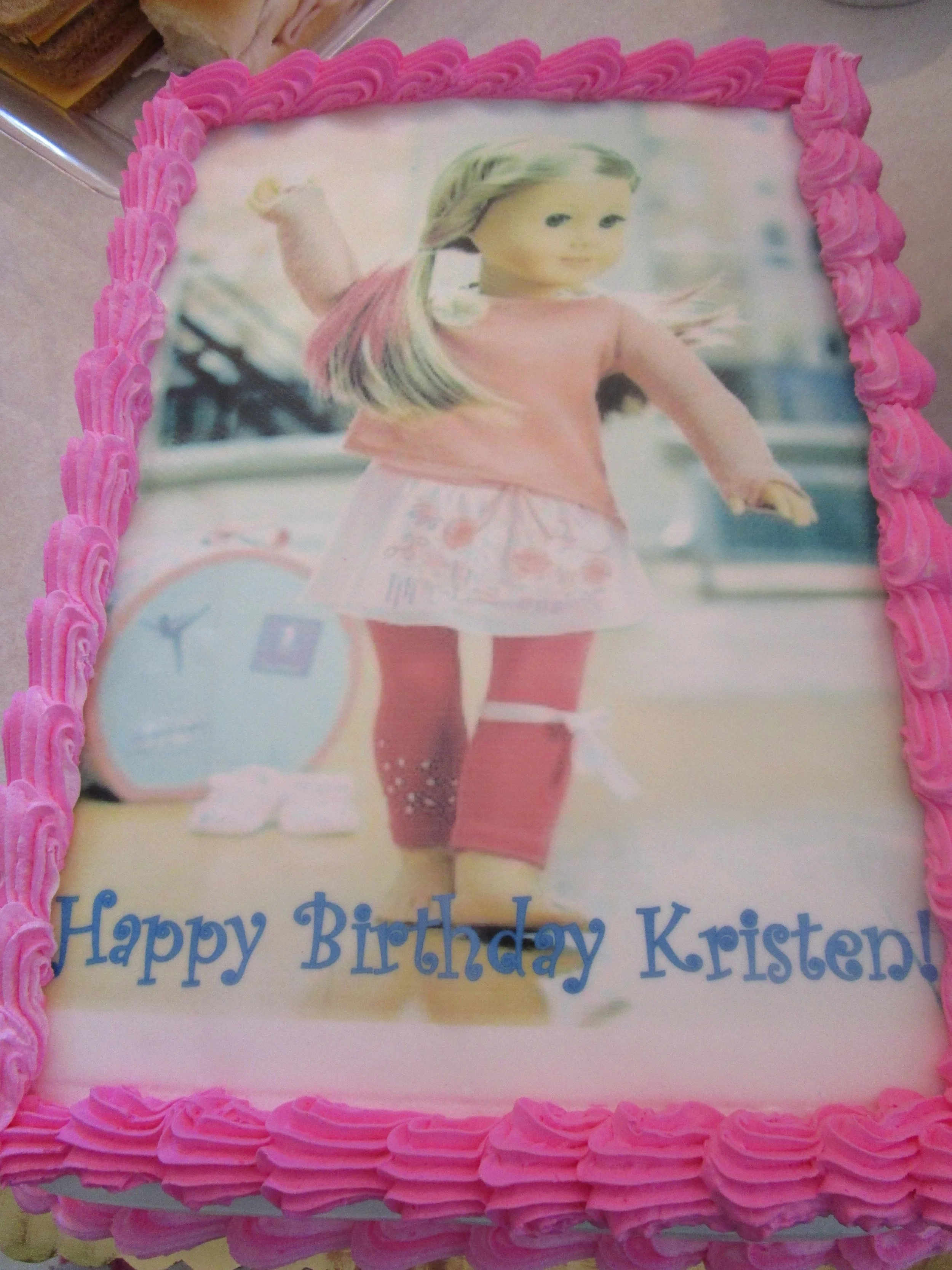 A birthday cake with a printed image of a young girl wearing pink clothing and standing outdoors, with pink frosting along the edges and the message "Happy Birthday Kristen!" written in blue letters.