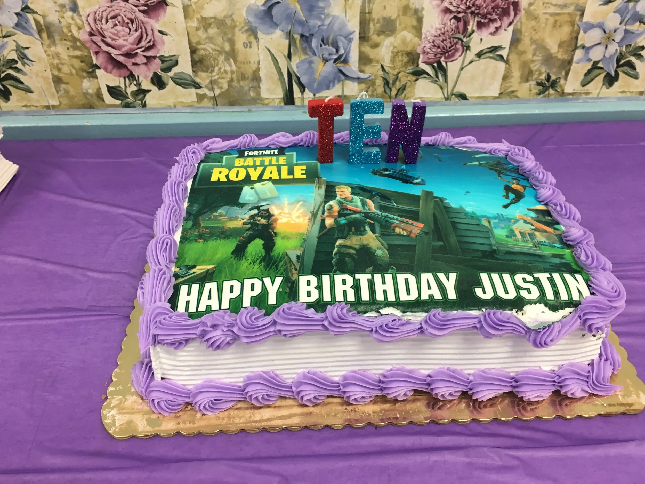 A birthday cake decorated with Fortnite Battle Royale theme, featuring purple and white icing and a picture of Fortnite characters. The cake has colorful glittered letters spelling T, E, and N on top, and a message that reads 'HAPPY BIRTHDAY JUSTIN'.