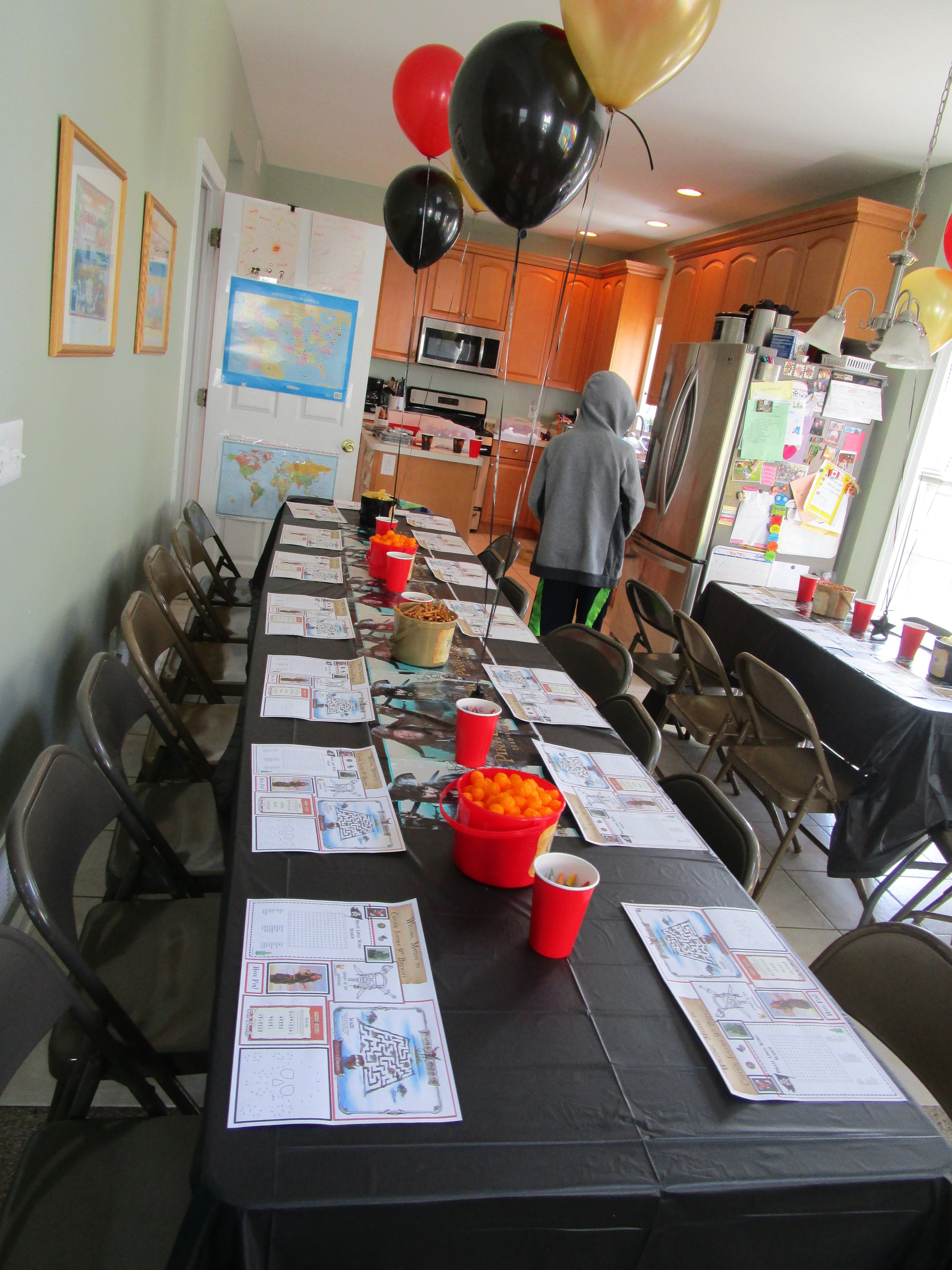 Long table set up for a party with papers, red cups, and bowls of snacks, black, gold, and red balloons floating above, in a kitchen with wooden cabinets and a refrigerator covered in magnets and papers, a person in a gray hoodie stands near the door.