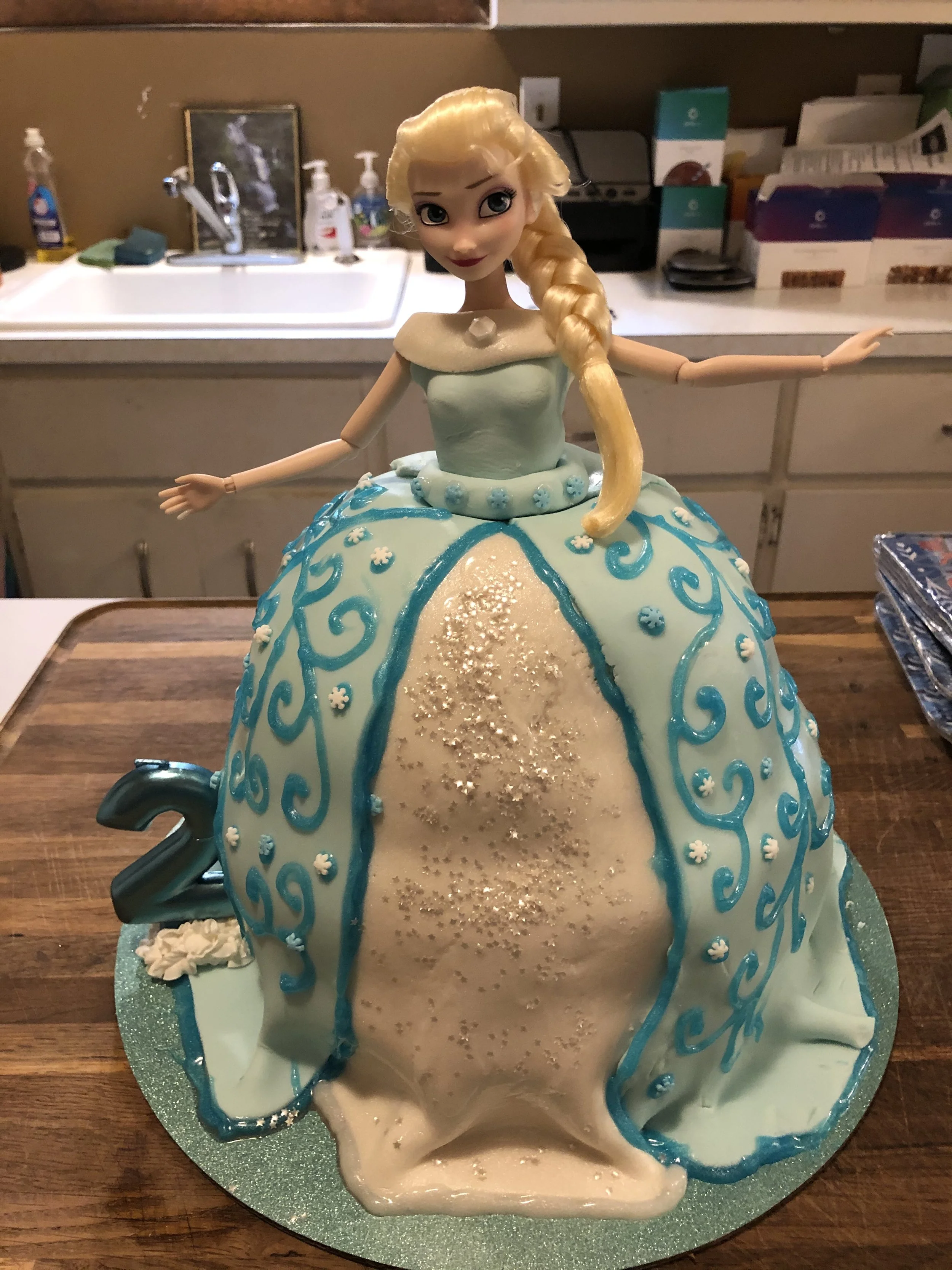 A cake shaped like a princess dress with a doll as the top, featuring blonde braided hair. The cake is decorated with blue swirls and white accents, includes a silver star and the number 2, suggesting it is for a second birthday celebration.