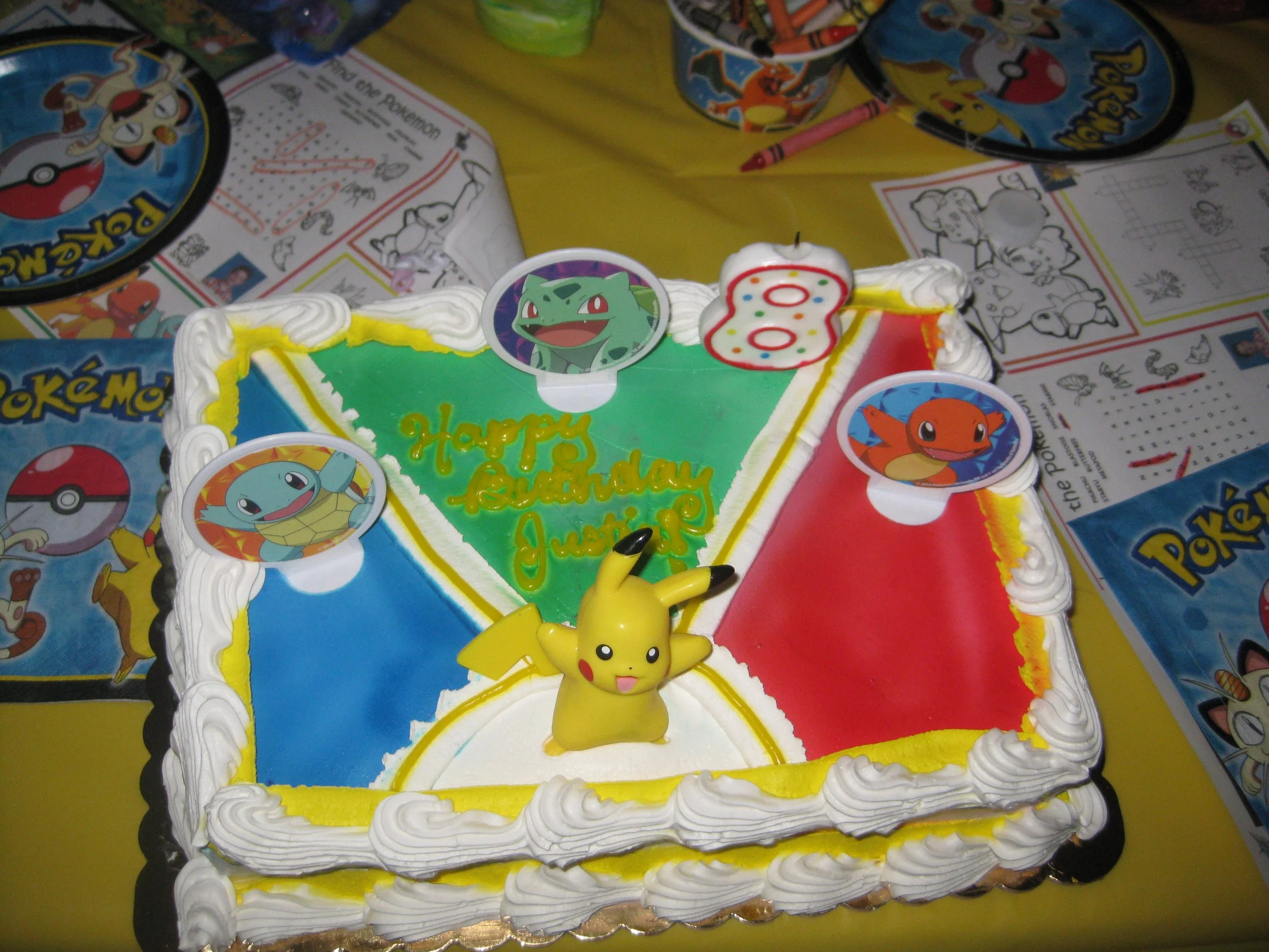 A colorful Pokémon-themed birthday cake with Pikachu, Bulbasaur, Charmander, and Squirtle decorations. The cake has sections in green, red, and blue, with little Pokémon stickers and a yellow Pikachu figurine on top. The table has Pokémon-themed supplies and coloring pages surrounding the cake.