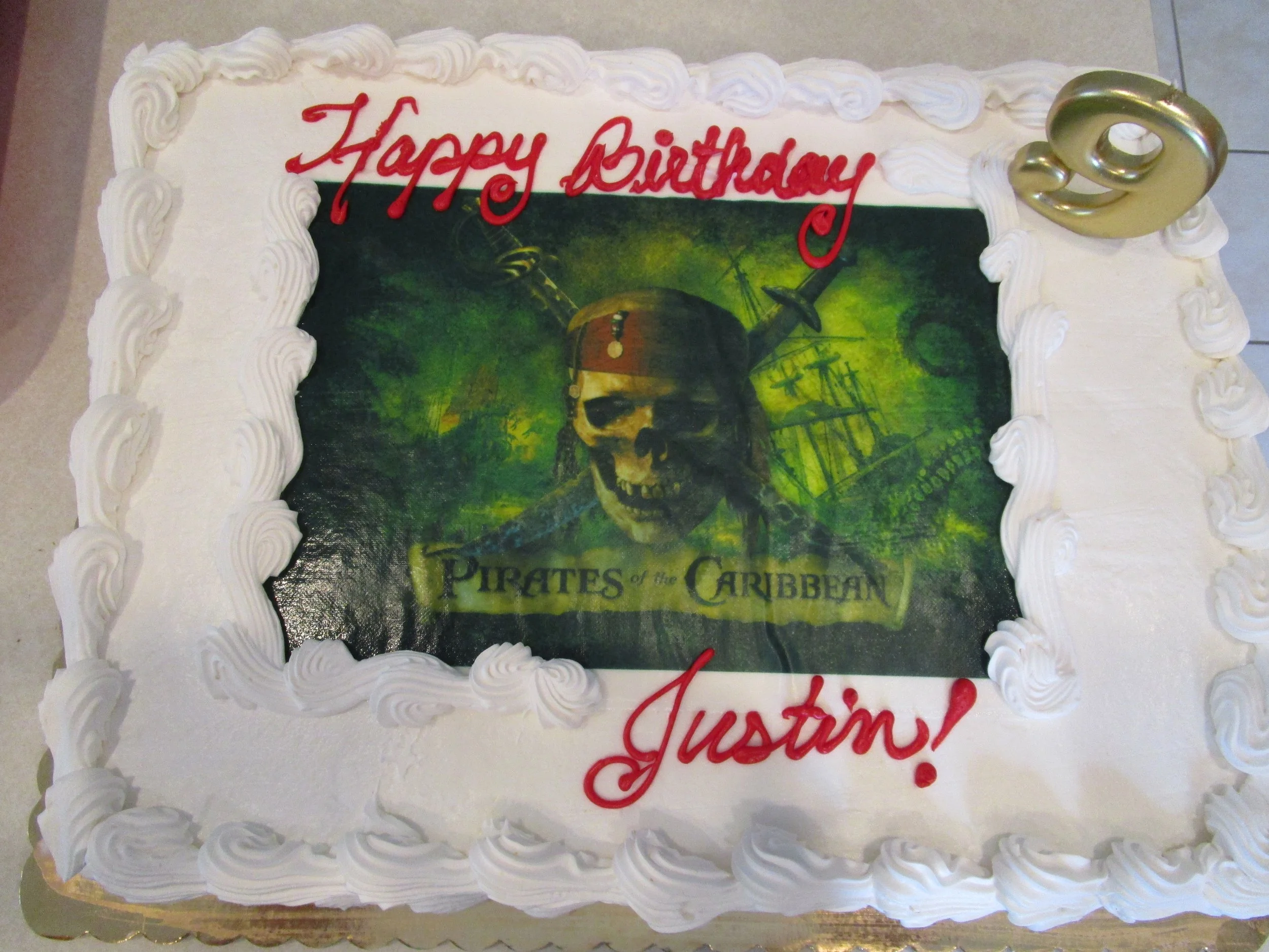 A birthday cake celebrating Justin's birthday with a decorated edible image of the Pirates of the Caribbean movie poster on top. The cake has white frosting with piped borders and a large gold number nine on the corner.