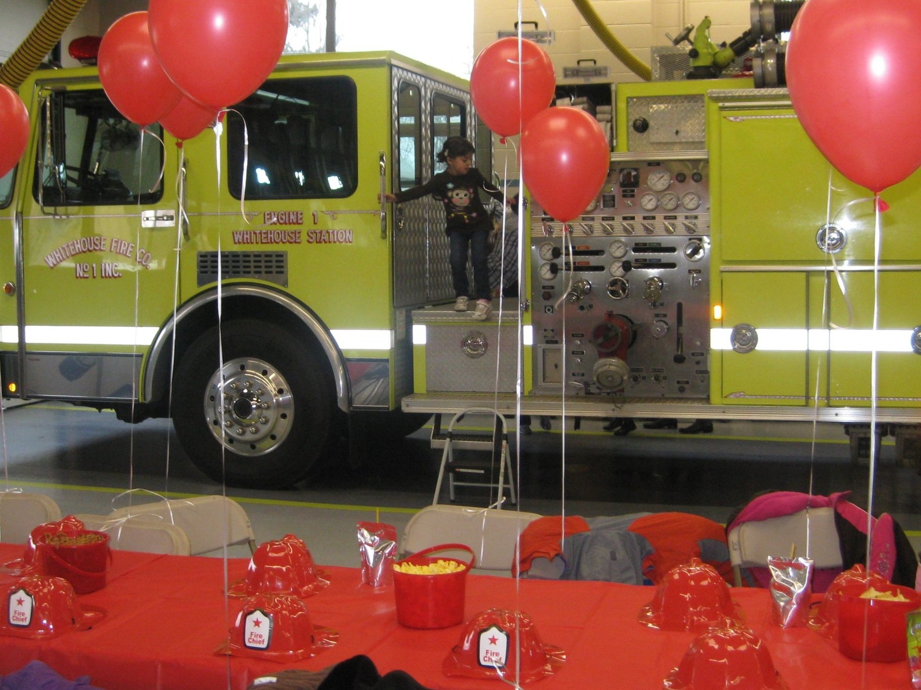 A young girl is climbing into a yellow fire truck during a celebration with red balloons and a red tablecloth decorated with fire hats and fire chief badges.