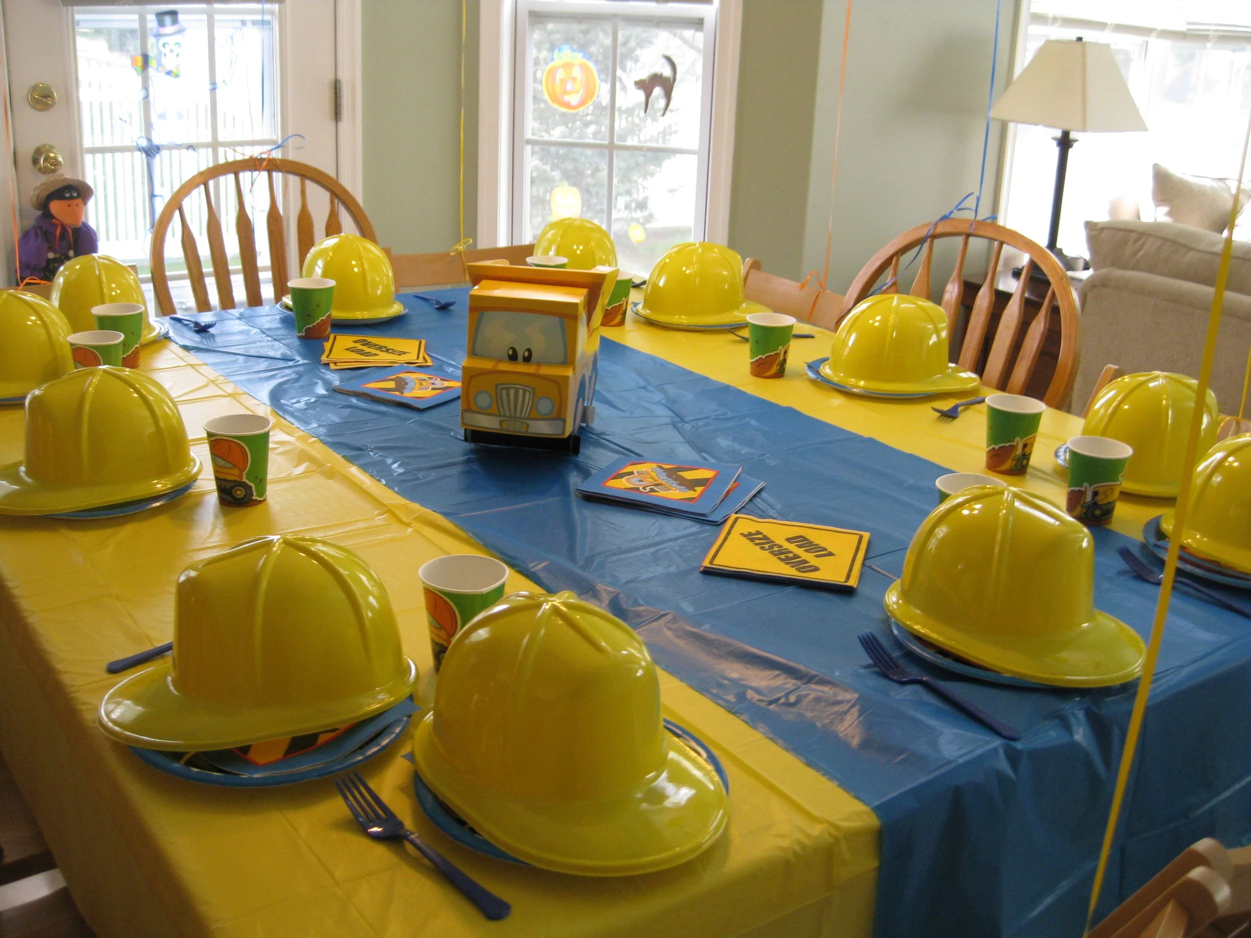 A party table decorated with construction theme items, including yellow plastic hard hats, paper cups with construction vehicle designs, and matching paper plates. The table has a blue table runner and yellow tablecloths, with a toy construction truck centerpiece and construction-themed clues or cards labeled 'construction zone'.