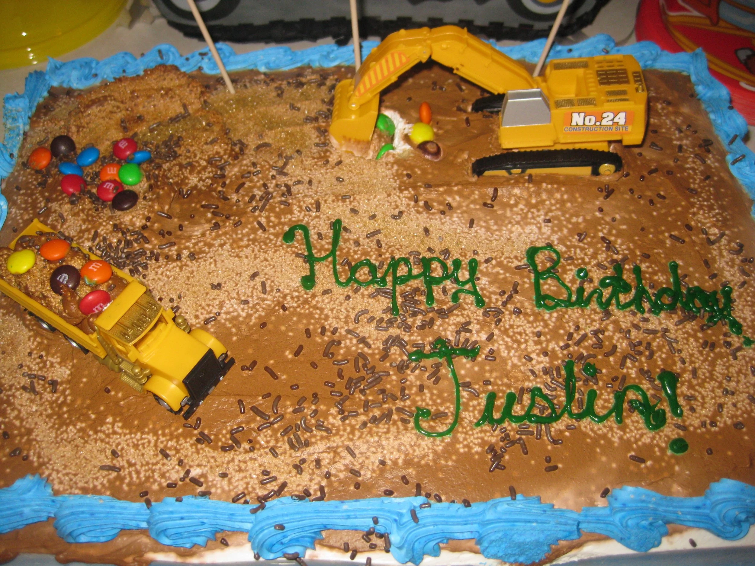 A birthday cake decorated with toy construction vehicles, colorful candies, and chocolate sprinkles, with green icing writing that says 'Happy Birthday Justice!'