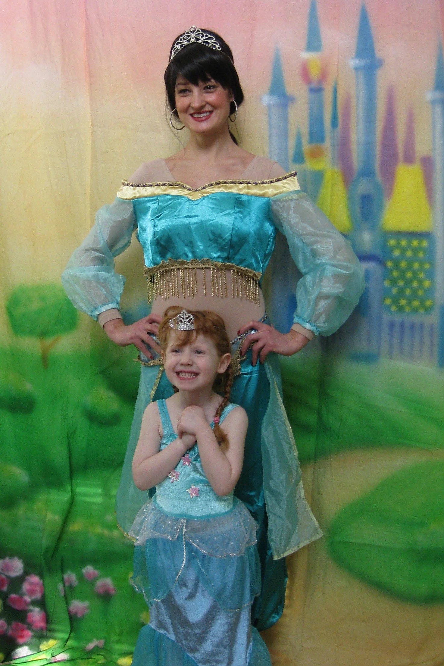 Woman dressed as a princess facing camera with a young girl in front of her, both smiling, in front of a colorful backdrop featuring a fairy tale castle and greenery.