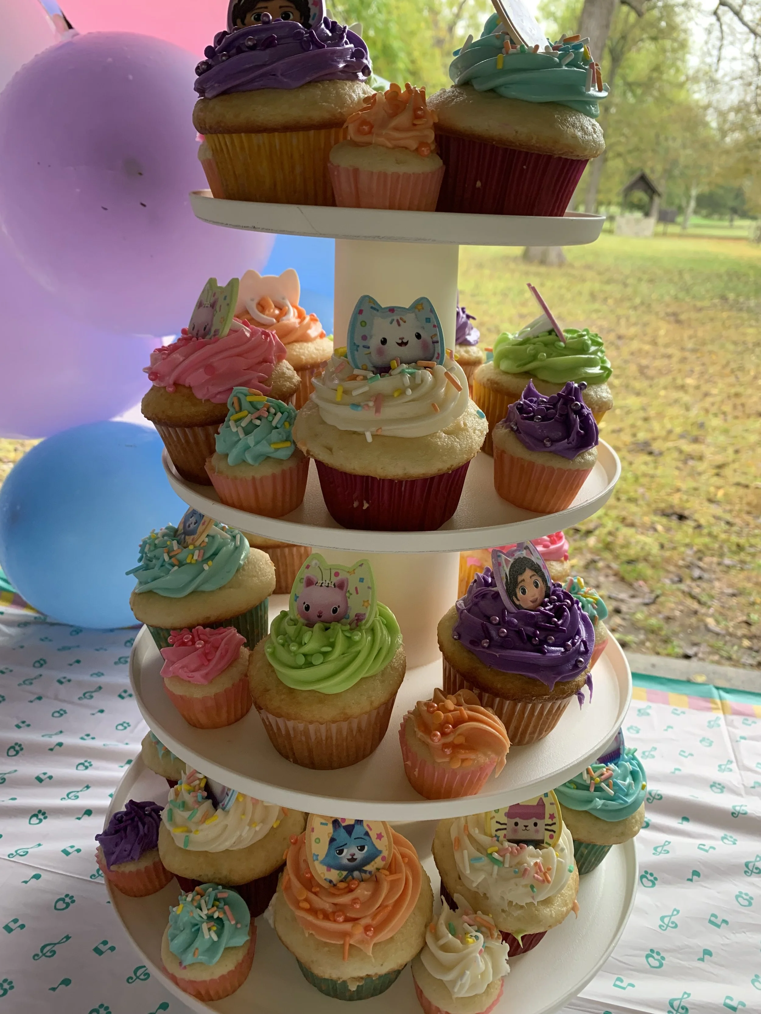 Colorful cupcakes decorated with rainbow sprinkles, frosting, and toppers featuring animated characters. The cupcakes are displayed on a white, multi-tiered stand with a background of trees and balloons.
