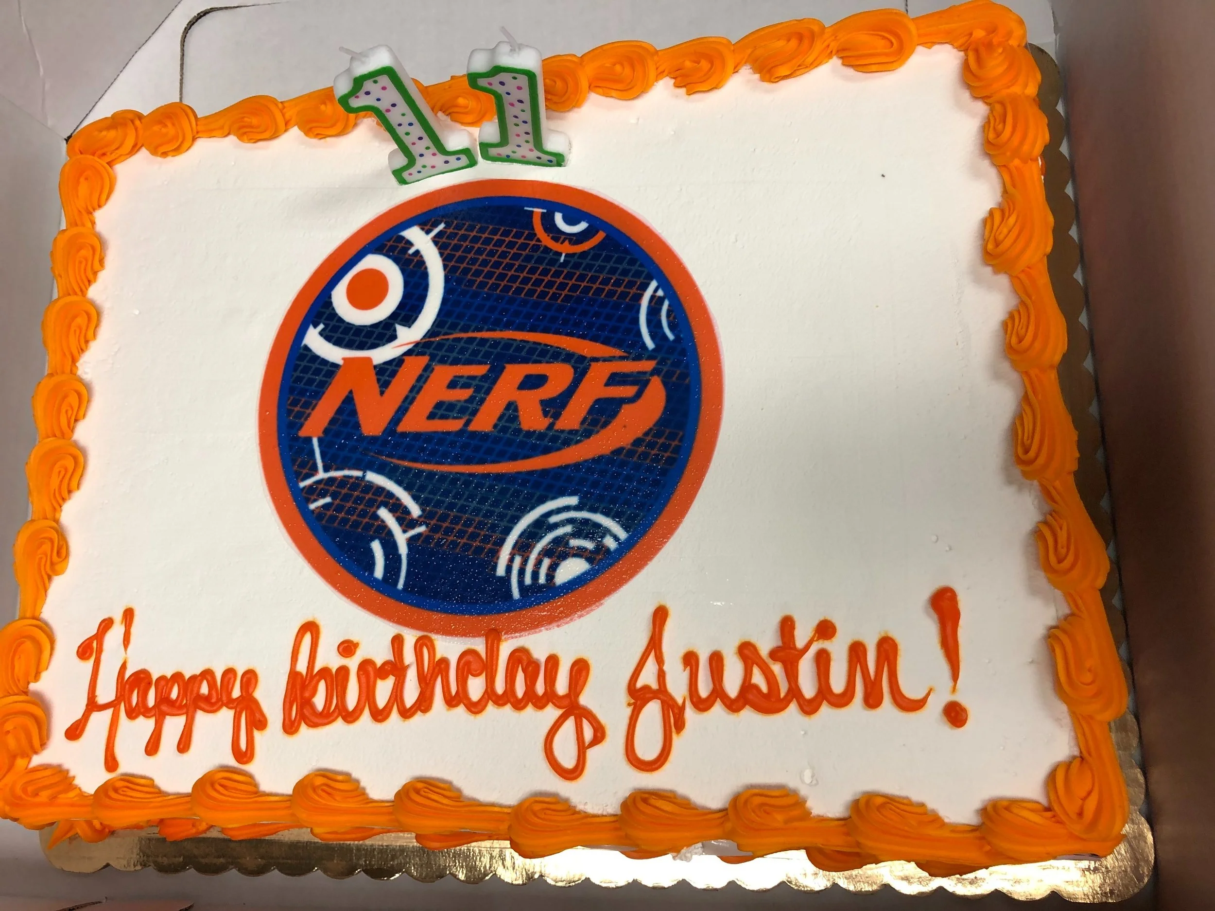A rectangular cake with white frosting, an orange border, and a Nerf logo in the center. There is a green and white '11' candle on top with the message 'Happy birthday Austin!' written in orange icing.