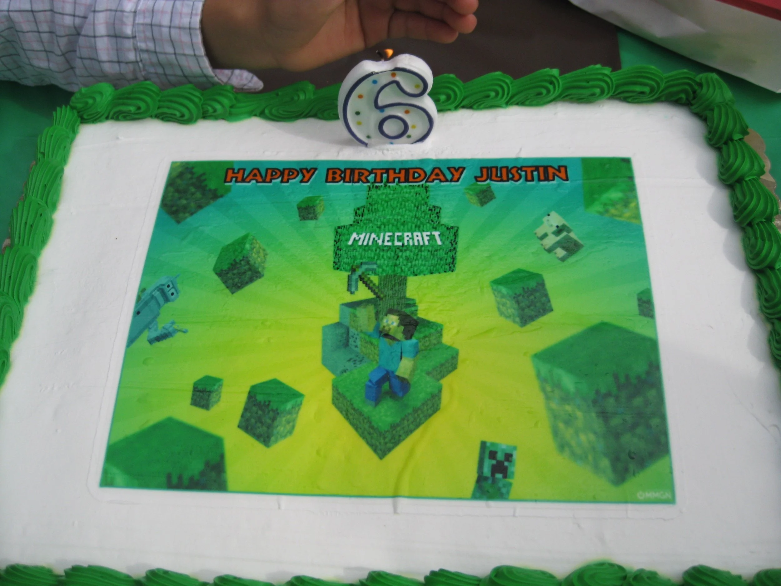 A birthday cake with a Minecraft themed edible image on top, surrounded by green frosting, with a decorated candle shaped like the number 6 on top. The cake appears to be celebrating Justin's sixth birthday.