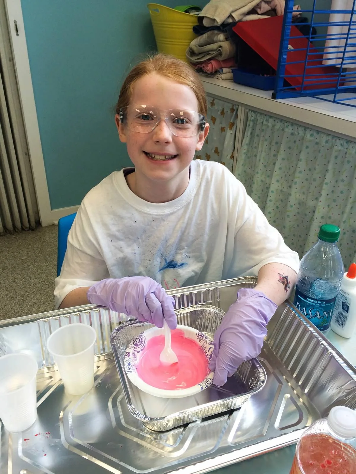 A smiling young girl with glasses and temporary tattoos on her arms is sitting at a table, stirring a pink and white mixture in a white bowl. She is wearing purple gloves and a white t-shirt. The table has supplies including cups, a water bottle, and a bottle of glue. In the background, there are shelves with folded clothes, a yellow bin, and colorful storage boxes.
