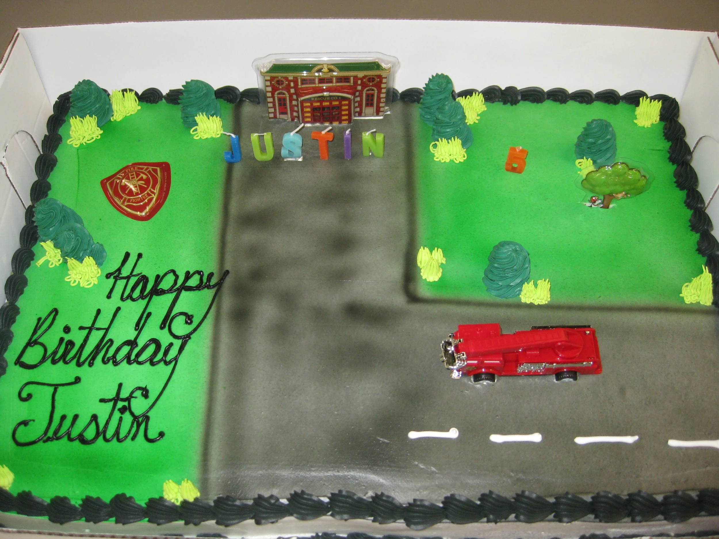 Birthday cake decorated with a fire truck, trees, a fire station, and colorful letters spelling 'Justin', with a message 'Happy Birthday Justin' on the cake.