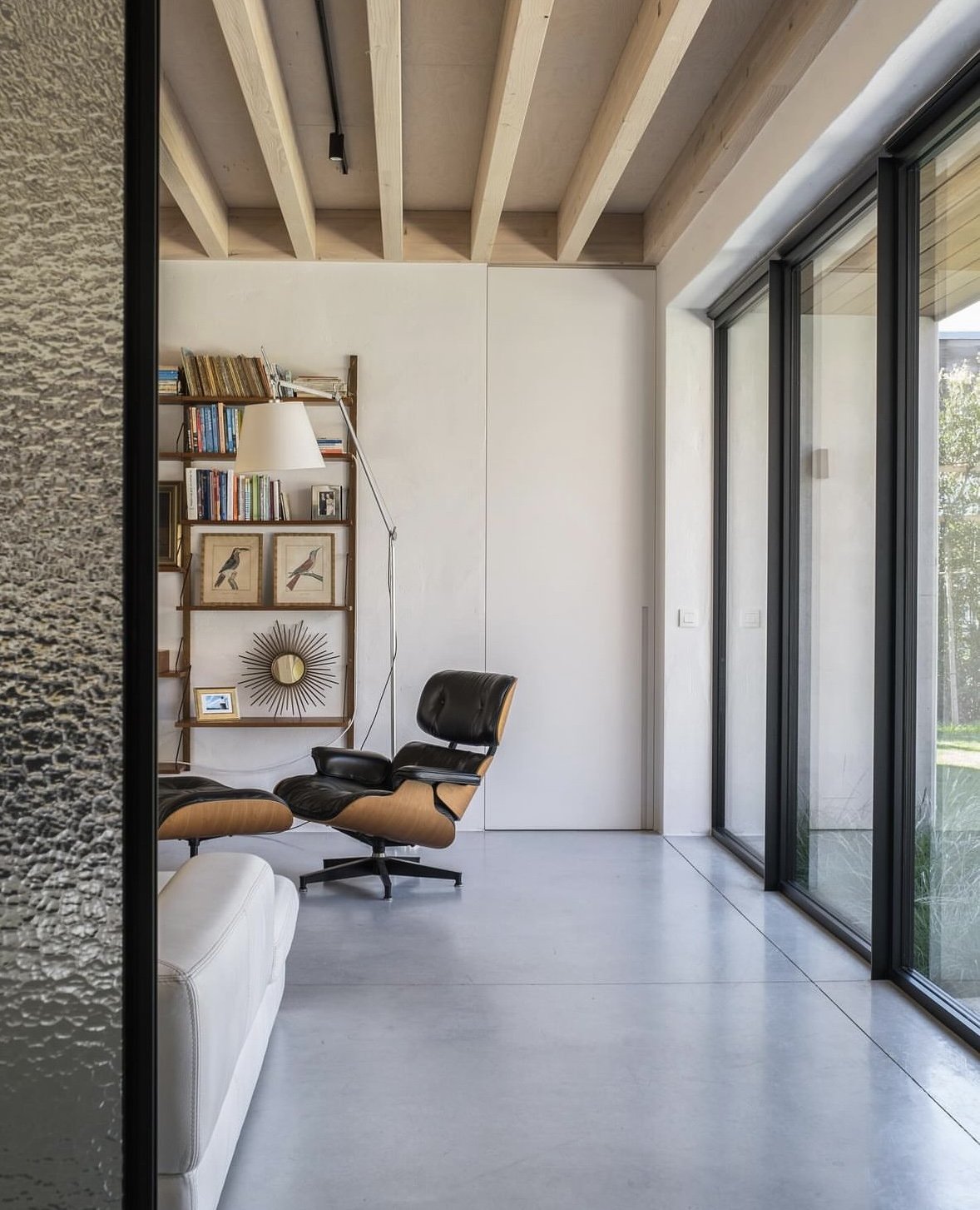 Modern living room with Eames lounge chair, tall bookshelf, large windows, and exposed wooden beams.