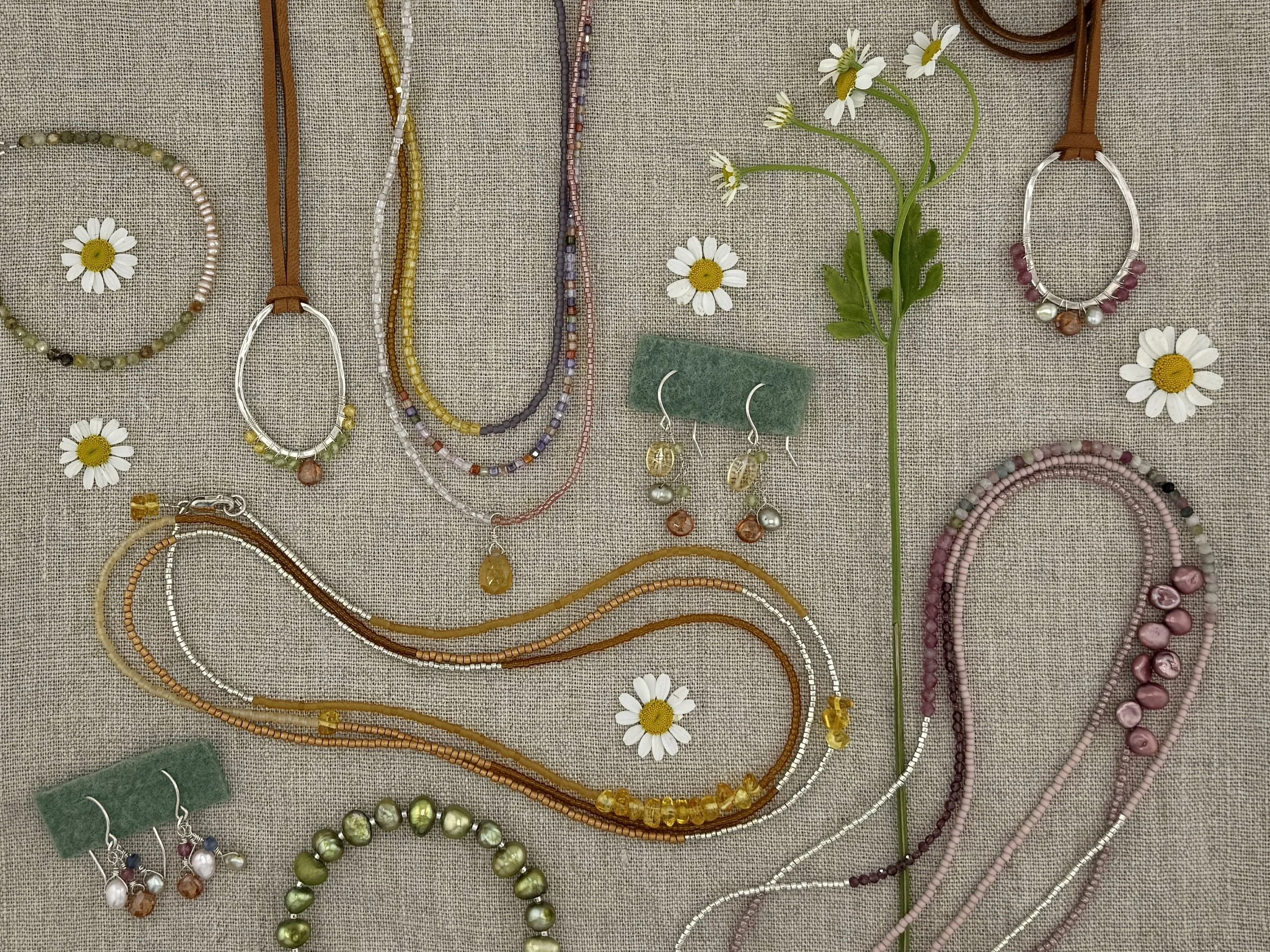 Assorted jewelry including necklaces, bracelets, earrings, and rings displayed on a beige fabric surface, decorated with small white daisies and a single wildflower.