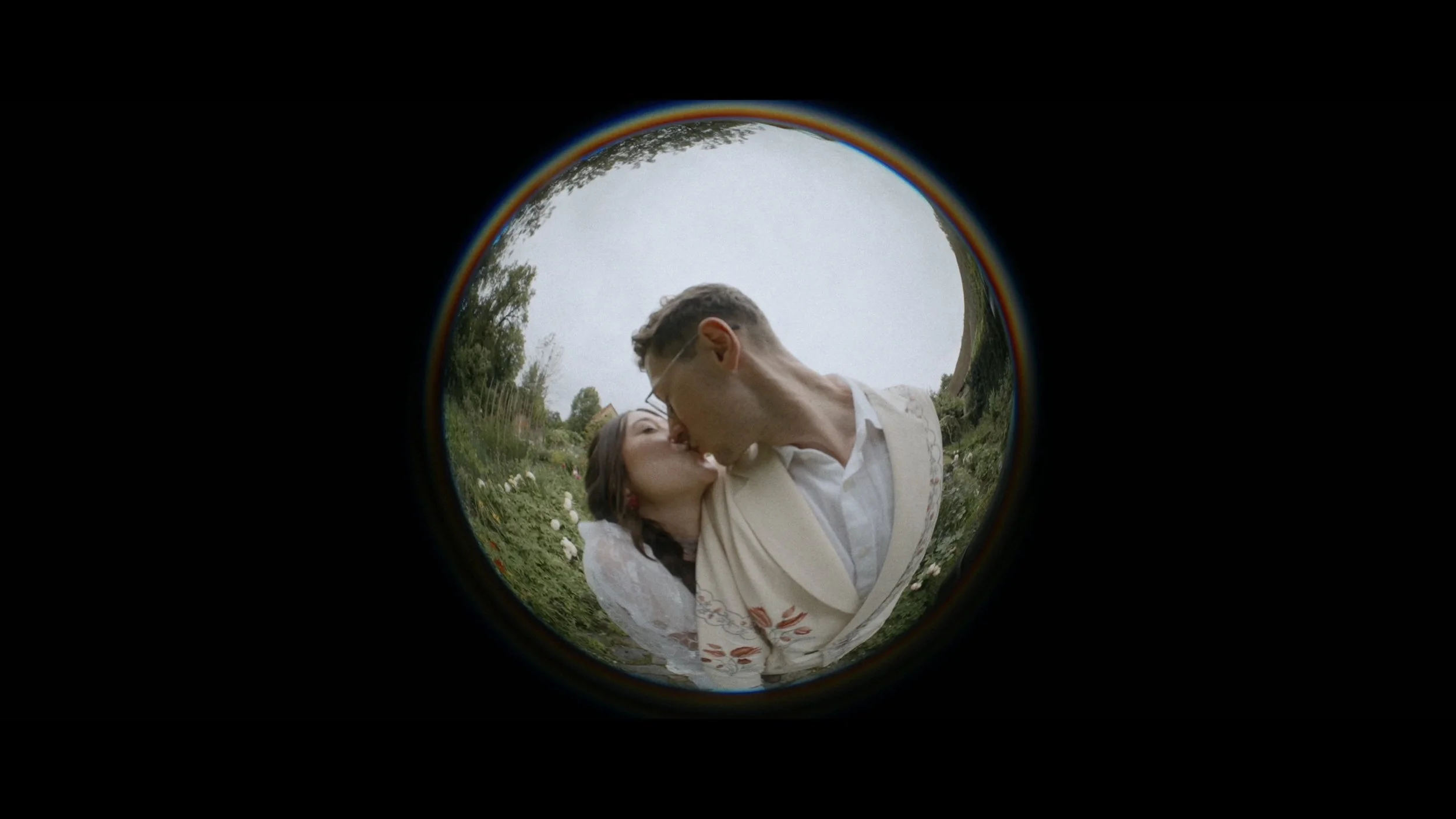A couple sharing a kiss seen through a fisheye lens, with greenery and floral garden in the background.
