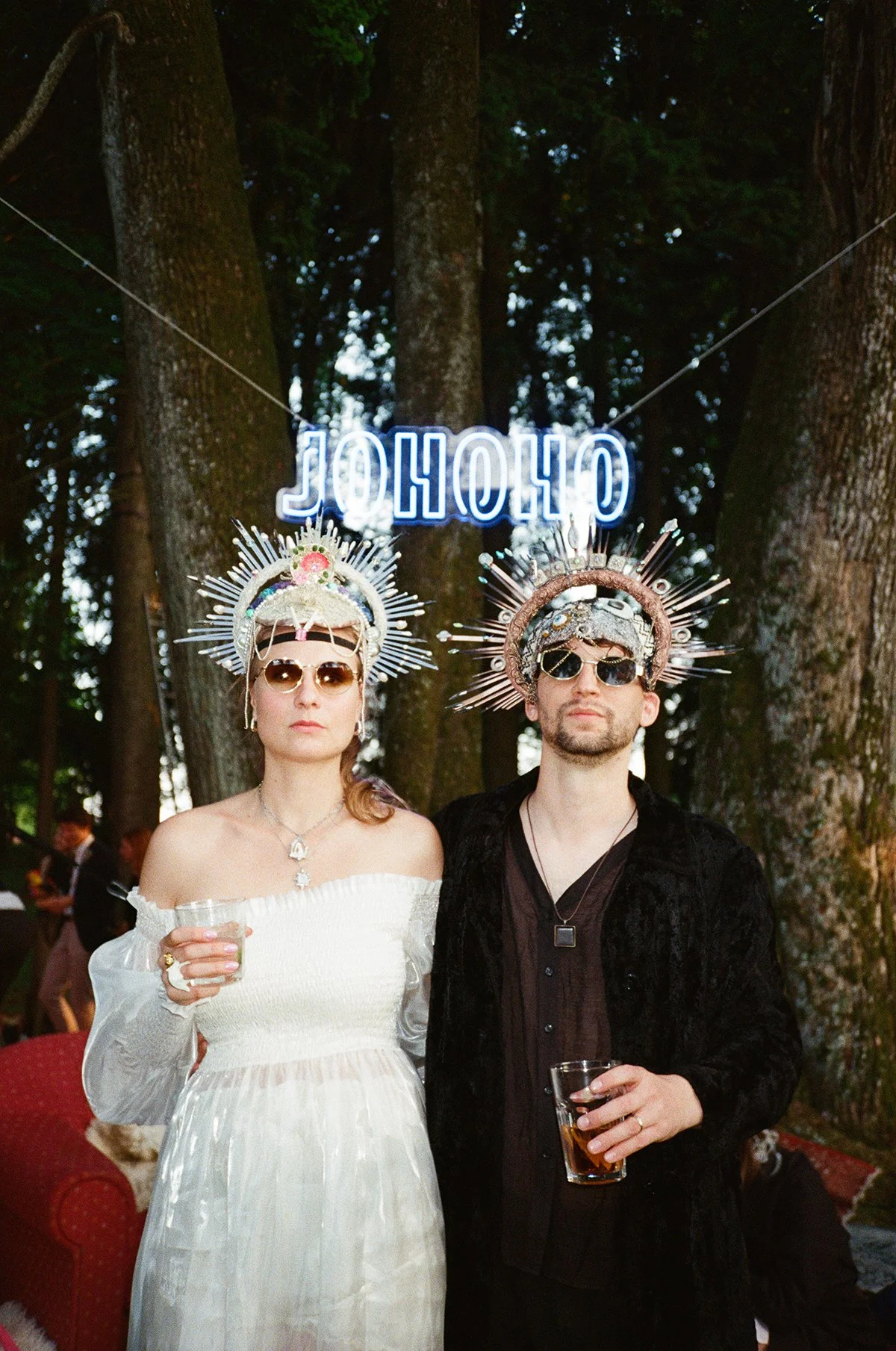 A woman and a man wearing ornate metallic crown-like headpieces and sunglasses, standing outdoors in front of a neon sign that spells 'OHOHO.' The woman wears a white off-the-shoulder dress and the man wears a black jacket and shirt, both holding drinks.