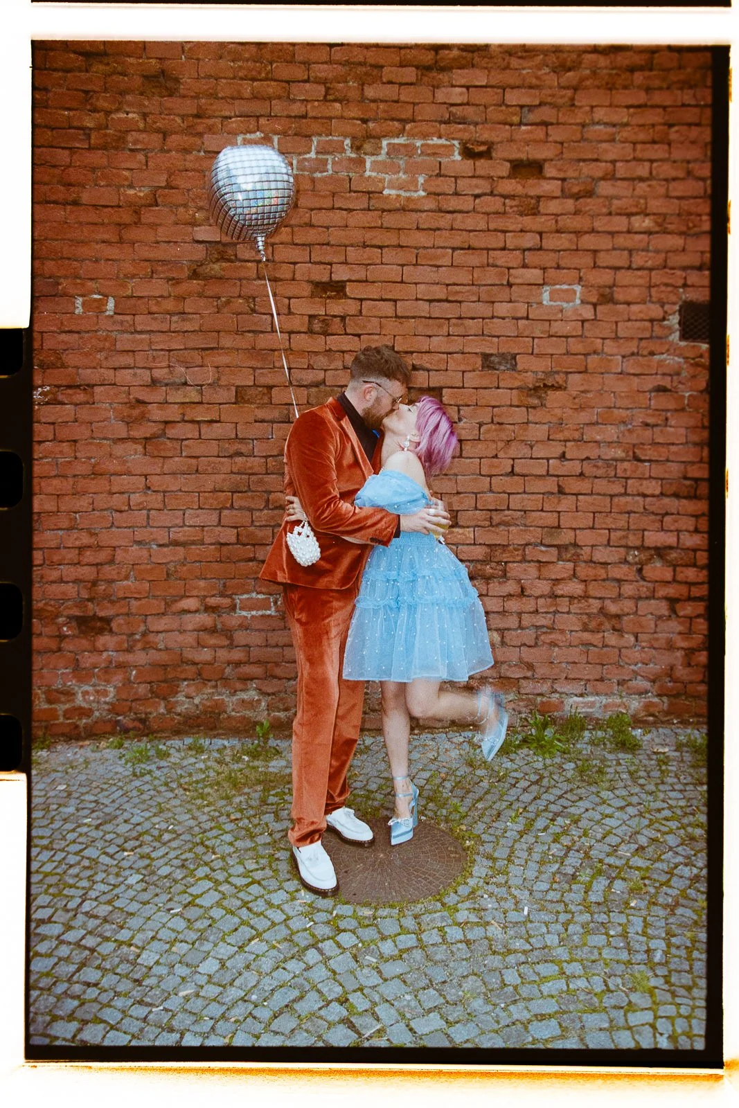 A couple sharing a kiss outdoors against a red brick wall, with the woman wearing a light blue dress and high heels, and the man in a rust-colored velvet suit holding a disco ball-shaped balloon.
