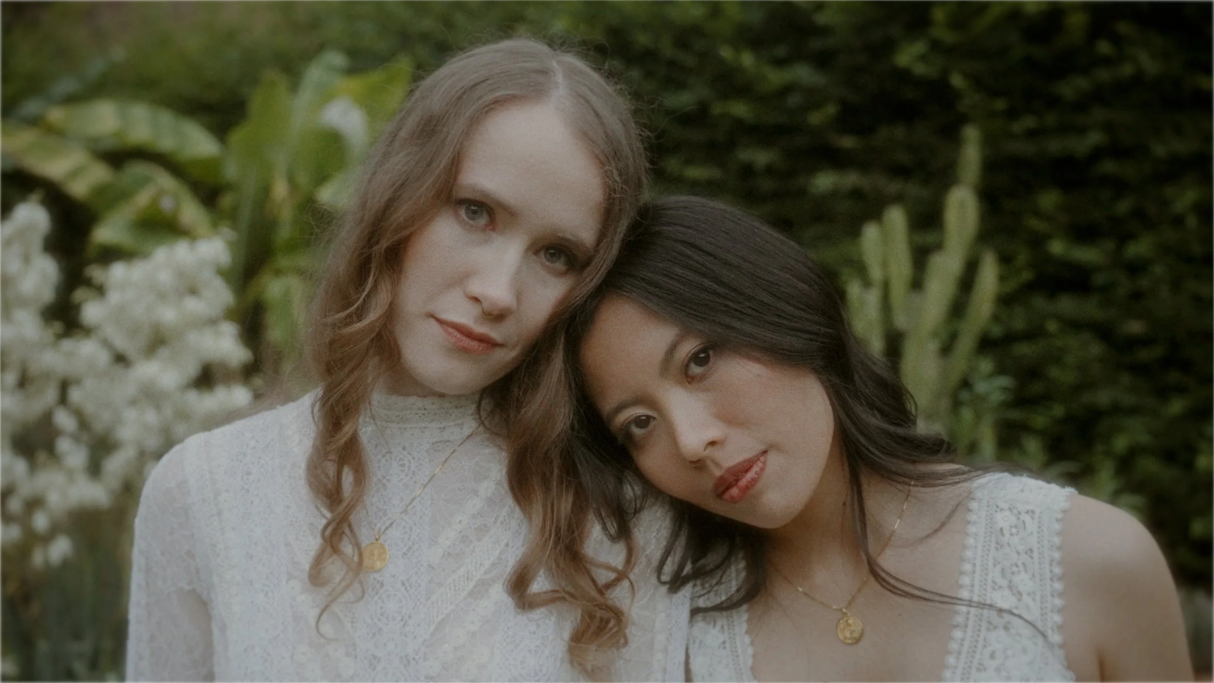 Two women standing close together outdoors, wearing white tops with lace details, each with a gold necklace, with greenery and cacti in the background.