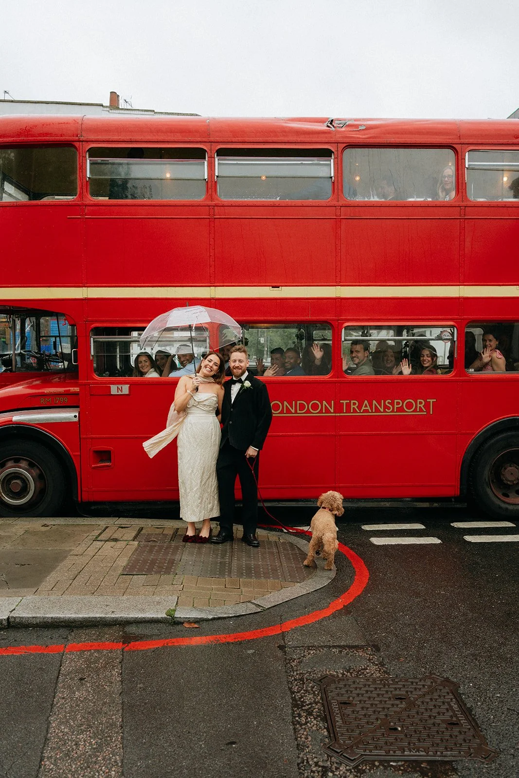 A newlywed couple stands in front of a red London double-decker bus, with the bride holding an umbrella and a small dog on a leash nearby. Passengers inside the bus are smiling and waving, celebrating.