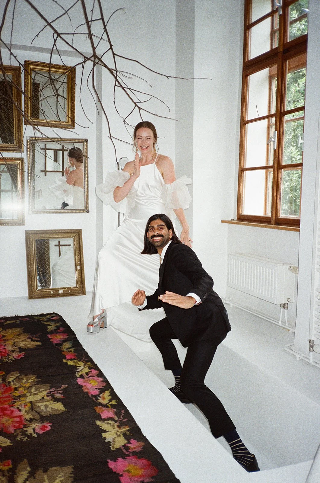 A woman in a white dress with puffed sleeves and high heels standing on a platform, smiling, while a man in a black suit crouches down near her, also smiling, inside a bright room with large windows, art on the walls, and a floral rug on the floor.