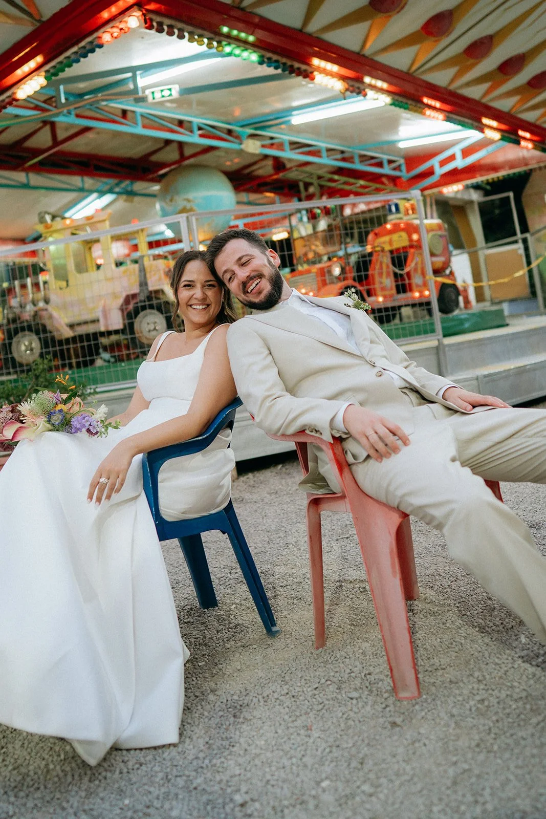 A newlywed couple sitting on colorful plastic chairs outside a funfair rides, smiling and enjoying their wedding celebration.