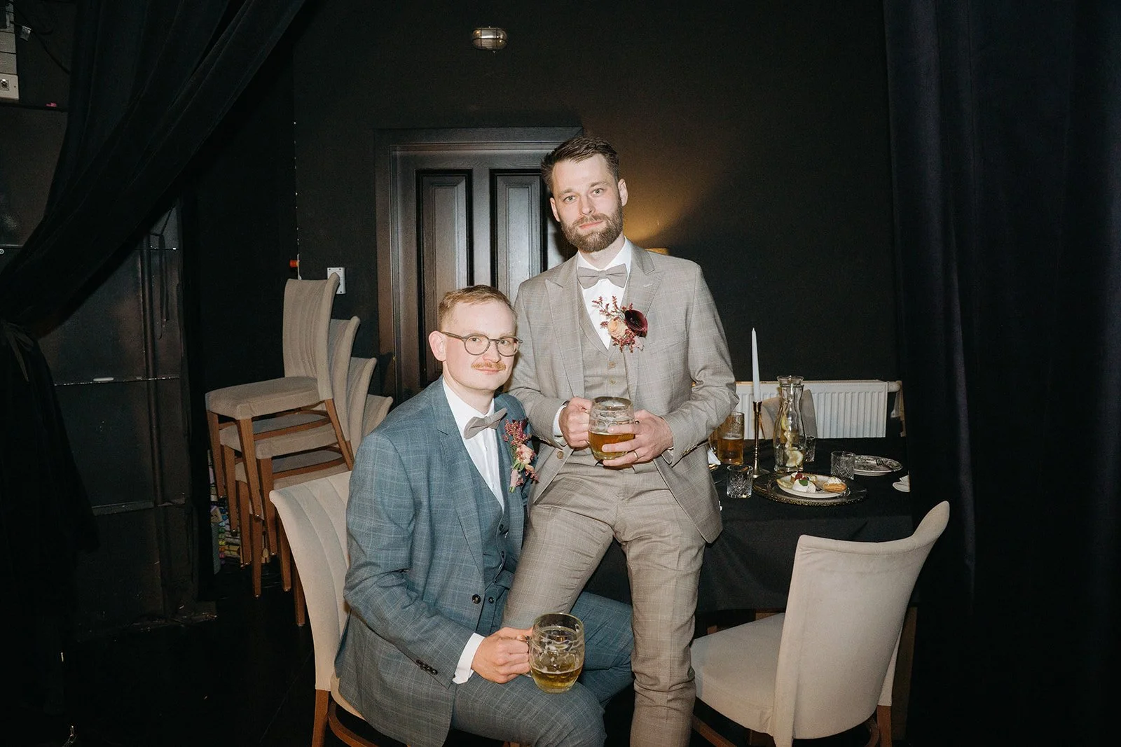 Two men in suits holding beer mugs at a formal gathering or wedding reception, standing in front of a decorated table with food and drinks.