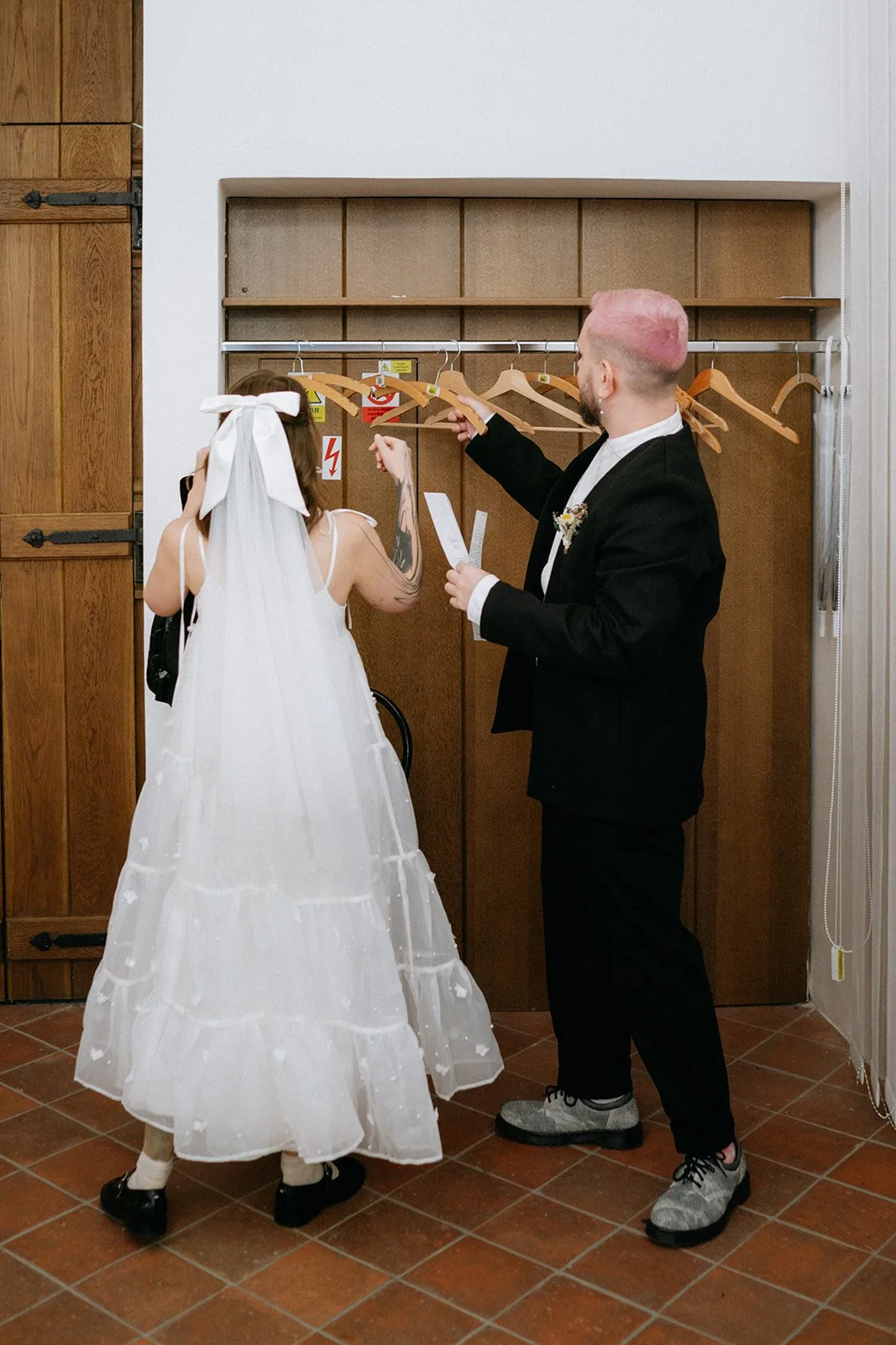 A bride and groom indoors, near a wooden wardrobe, with the groom holding a piece of paper and reaching for a wooden hanger, while the bride, dressed in a white gown with a large bow in her hair, looks at the hangers.
