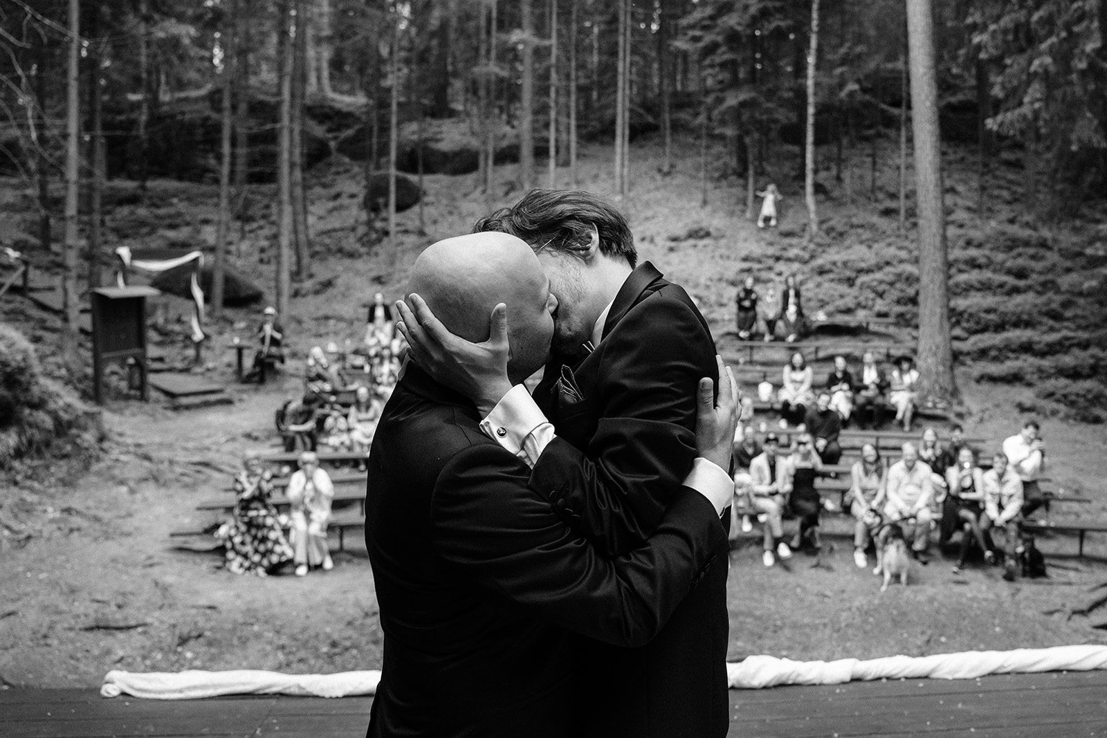 Two men in tuxedos kissing and embracing at an outdoor wedding ceremony in a wooded area.