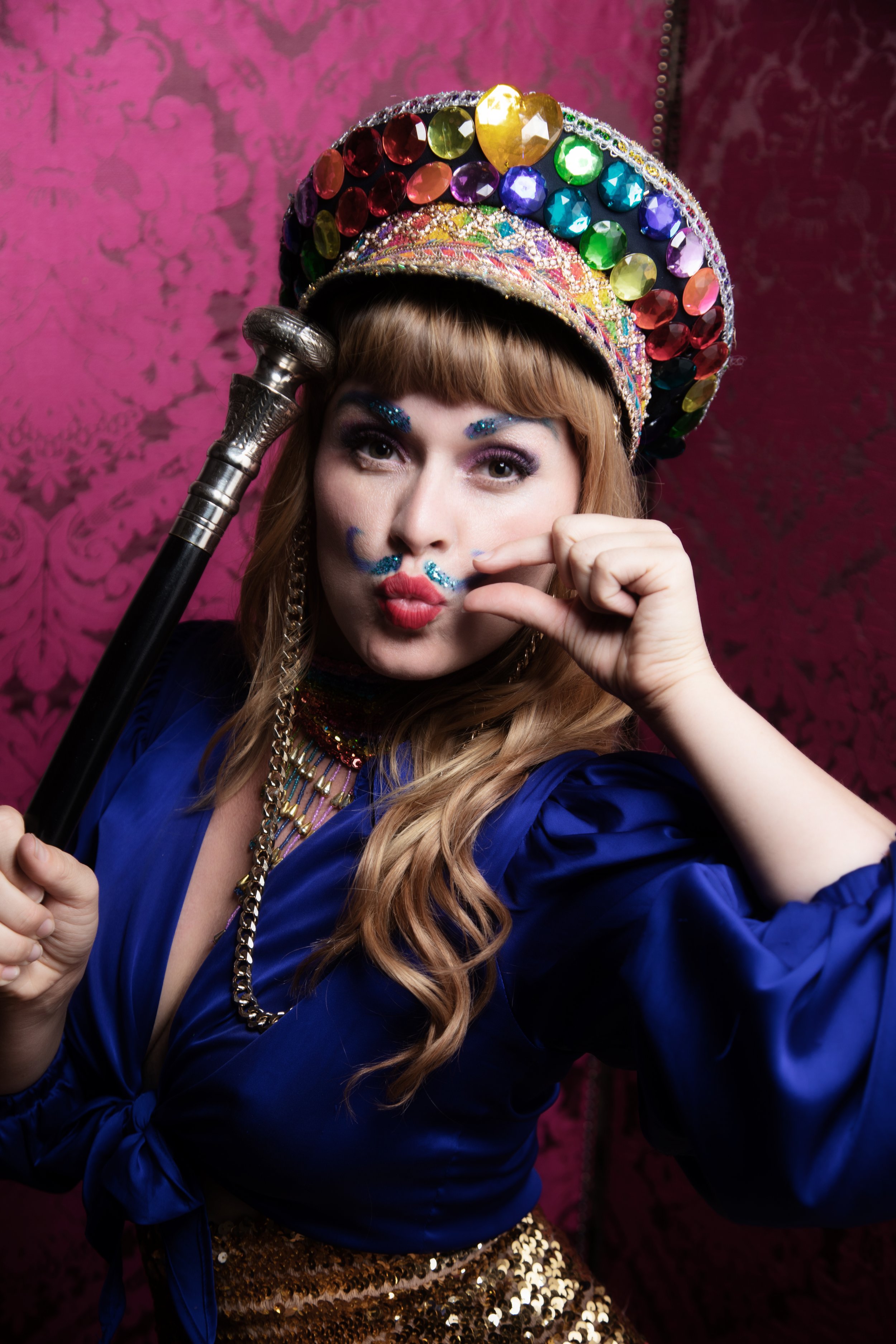 A burlesque performer in a brightly coloured, sparkly, hat holding a cane and wearing a fake glittery moustache and a blue shirt.