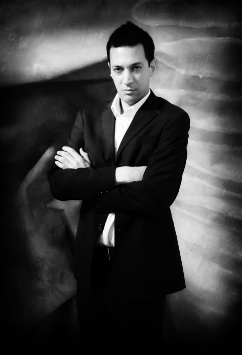 A man stands with his arms crossed, in a suit, in front of a x-ray backdrop.