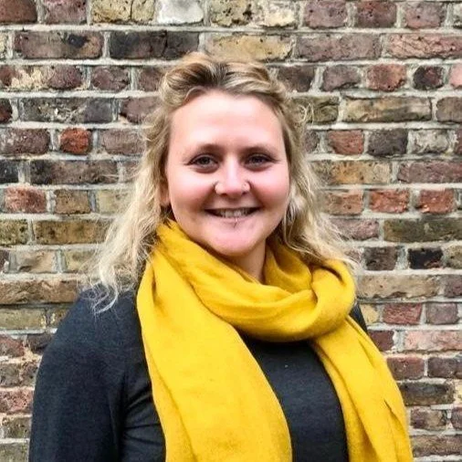 A photograph of Jami Dixon, Bright Impact CIC's founder.  Jami is wearing a bright yellow scarf, a black jumper and has blonde hair that she is wearing down.  Jami is smiling.