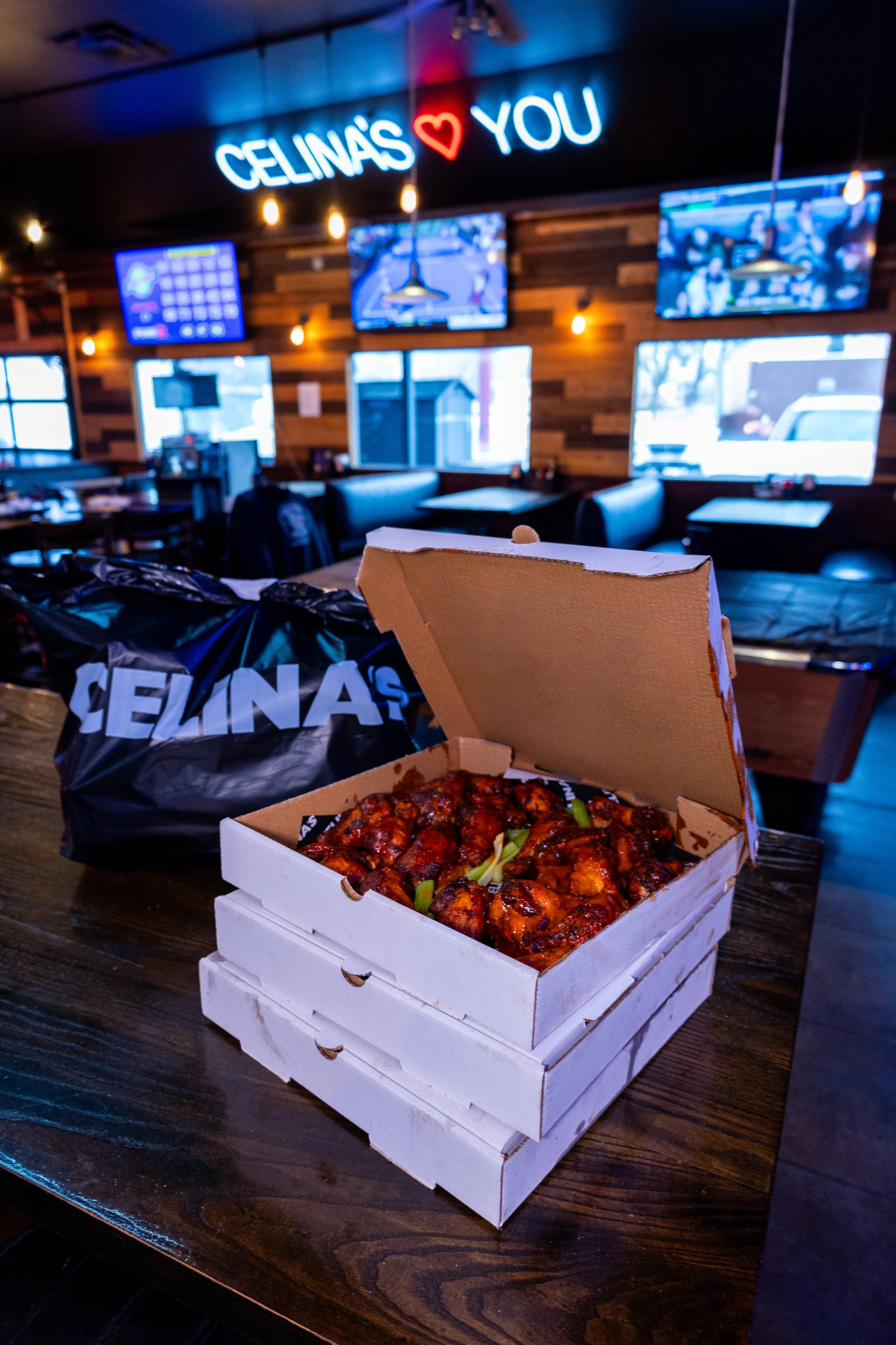 Pizza box containing takeout chicken wings with green onions inside, on a wooden table in a restaurant with TVs on the wall and a neon sign that reads 'CELINA'S ♥ YOU' in the background.