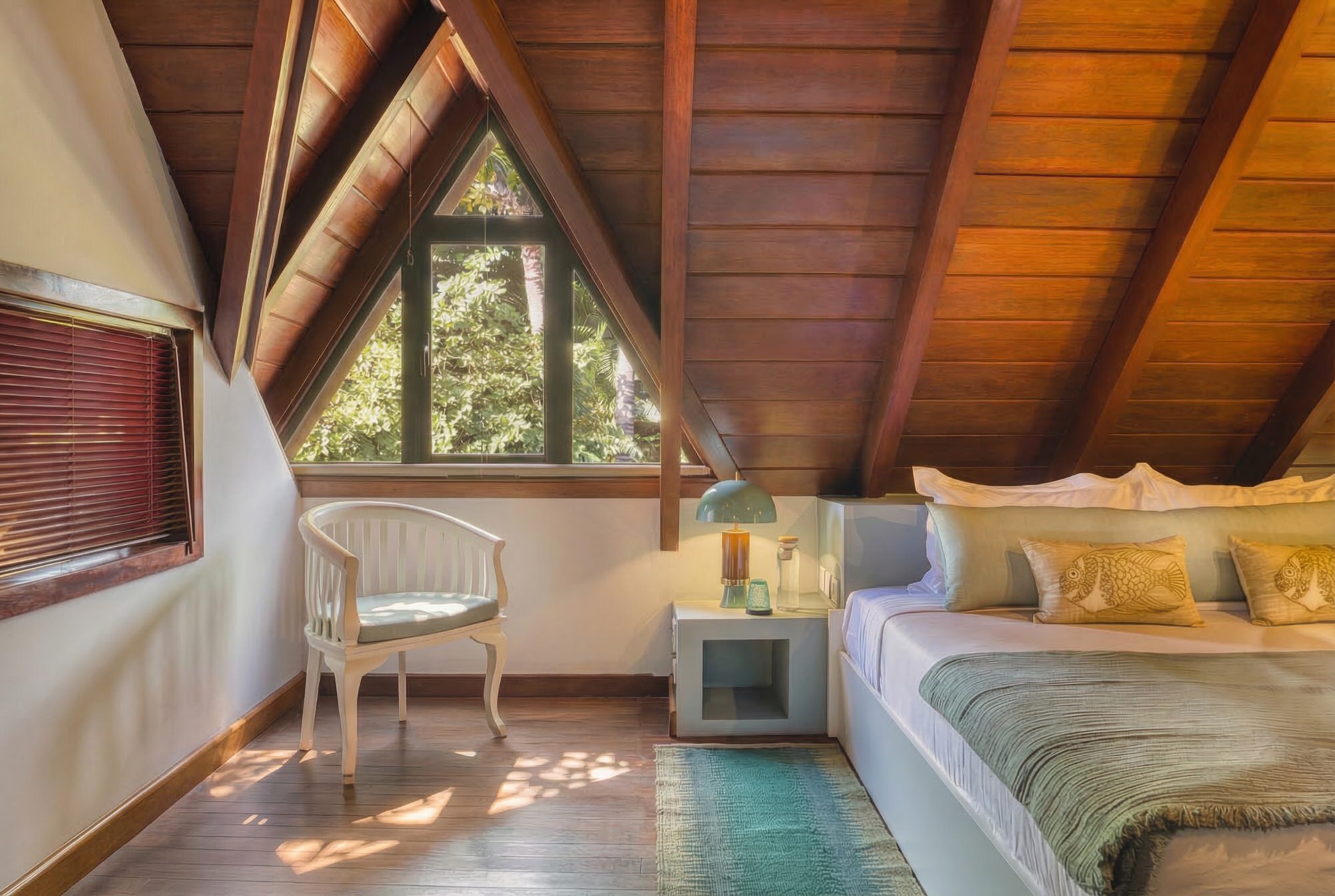 Chambre avec plafond en bois, grande fenêtre triangulaire laissant entrer la lumière et vue sur la verdure, lit avec coussins, lampe de table sur une table de nuit, chaise blanche près du mur