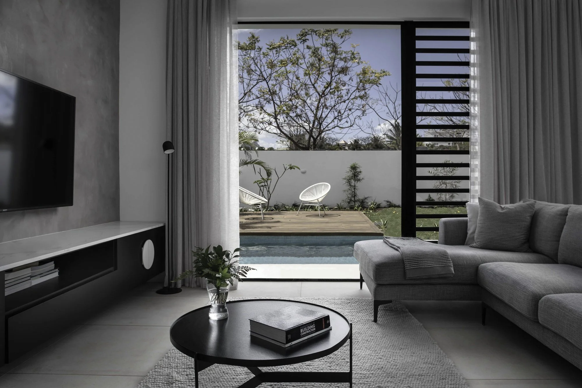 Intérieur moderne d'un salon avec une vue sur une terrasse extérieure, comprenant un canapé gris, une table basse ronde noire avec des livres, une télévision à gauche, des rideaux blancs, un arbre visible par la grande fenêtre, et des chaises en plas