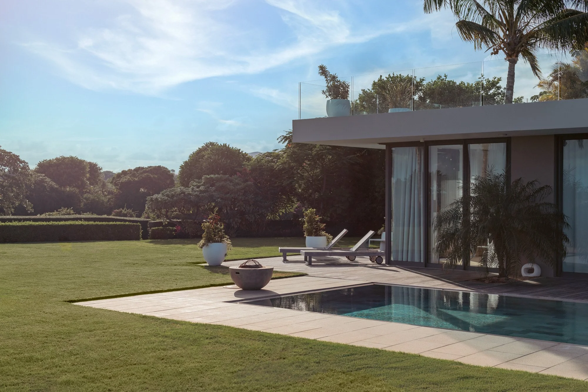 Vue d'une maison moderne avec une piscine, un salon de jardin, des chaises longues, et un jardin bien entretenu sous un ciel en partie nuageux, entourée de végétation tropicale.