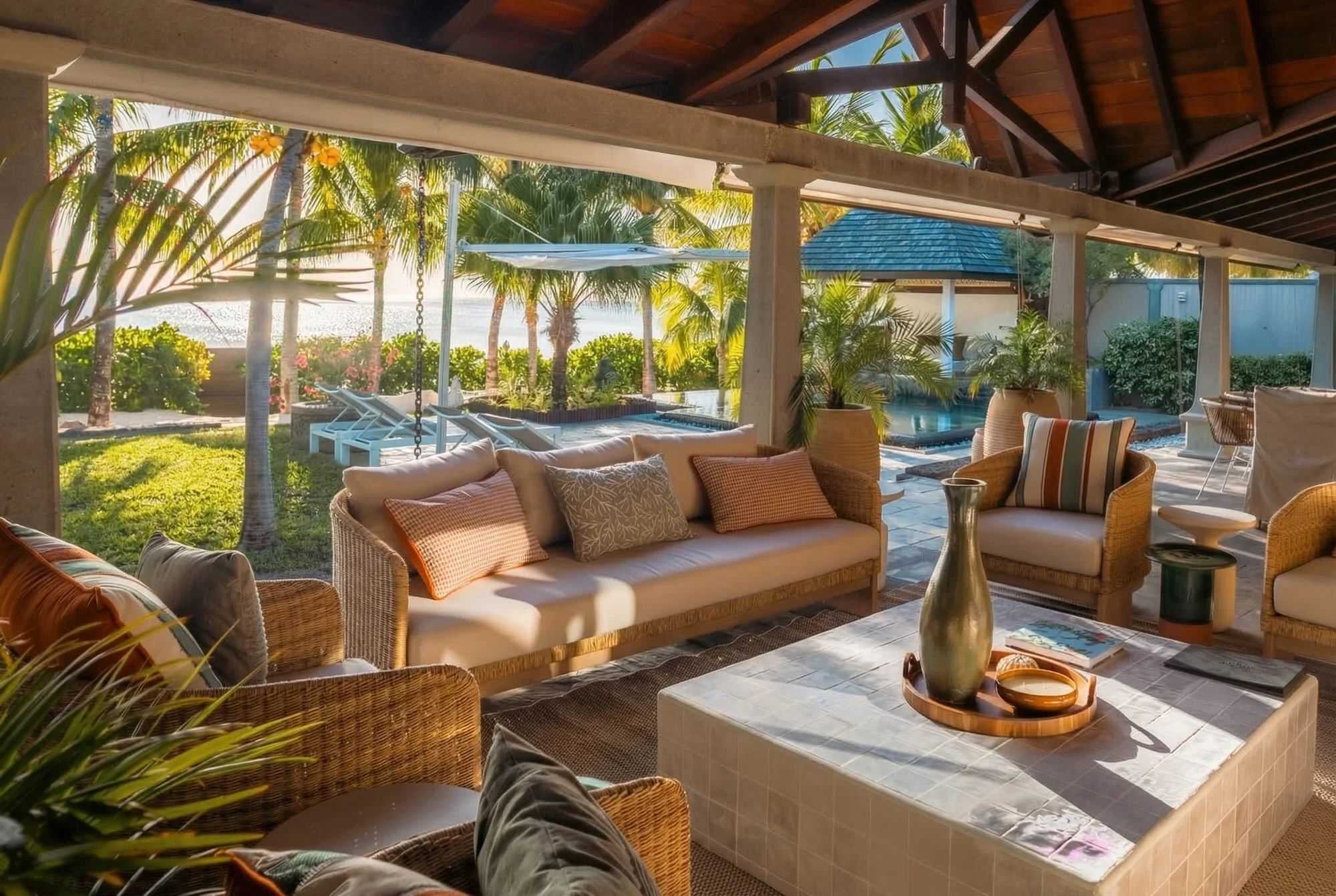 Terrasse ouverte avec mobilier en rotin, coussins et table basse, vue sur un jardin tropical avec palmiers, piscine et parasol, au bord de la mer.