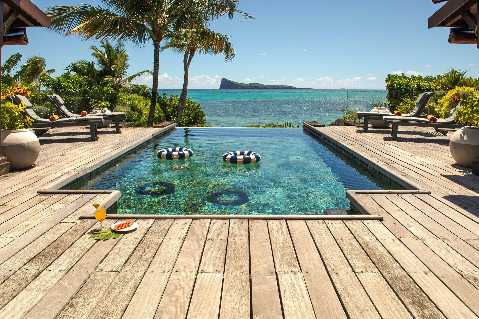 Terrasse en bois avec une piscine à débordement donnant sur la mer, avec des transats, des palmier et un paysage d'île rocheuse au loin, sous un ciel ensoleillé.