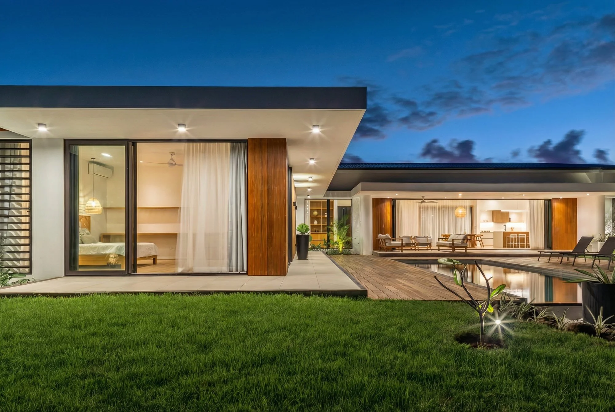 Maison moderne avec intérieur visible, grande terrasse en bois, piscine, et jardin bien entretenu, éclairée en soirée.