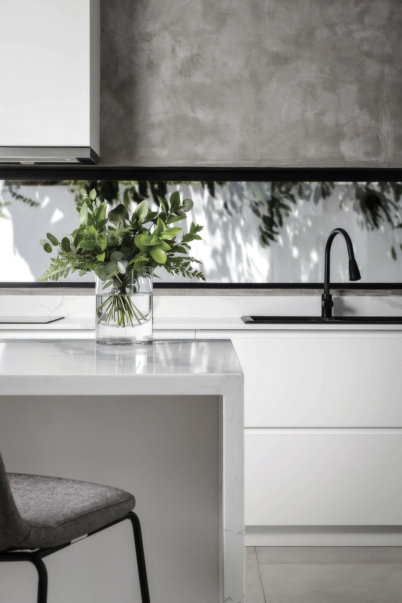 Une cuisine moderne avec un plan de travail blanc en marbre, un évier noir, un robinet noir, et une fenêtre avec un reflet de plantes vertes, une plante dans un vase en verre sur le comptoir.