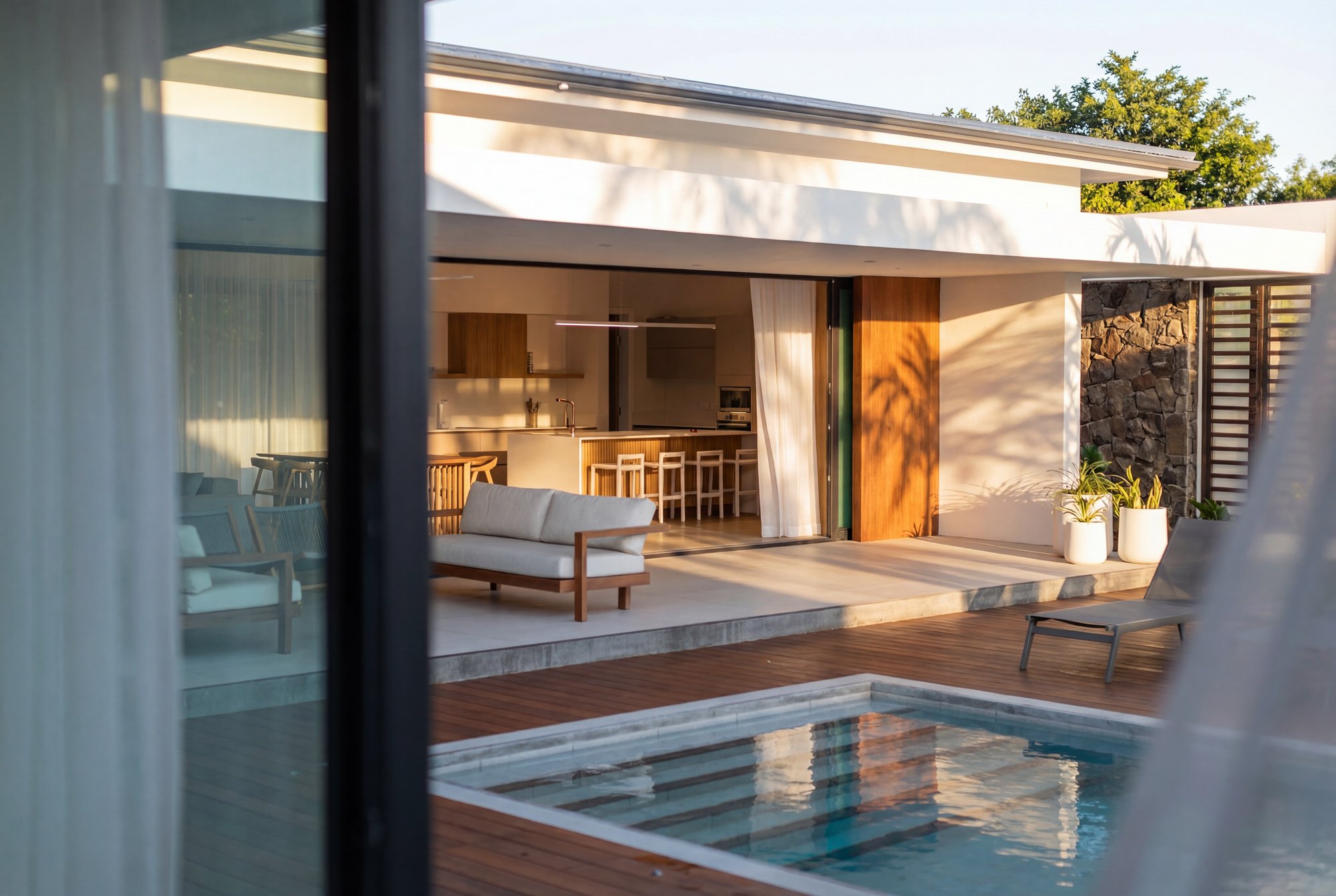 Vue depuis une fenêtre montrant un espace extérieur moderne avec piscine, mobilier en bois et plantes dans des pots blancs, maison en arrière-plan avec mur en pierre et jardin avec arbres.