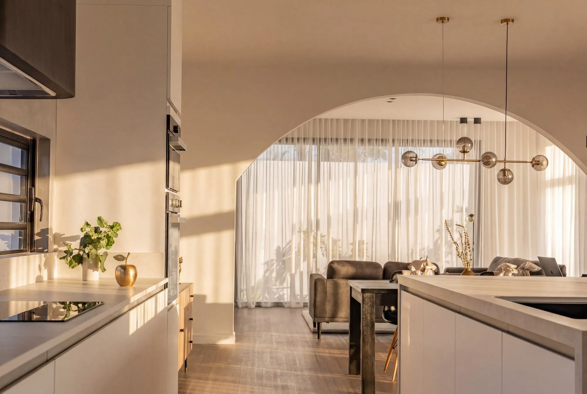 Intérieur moderne d'une cuisine ouverte avec un salon, éclairée par la lumière naturelle, comprenant un mobilier blanc, un comptoir en bois, une suspension avec des globes en verre, et des rideaux légers derrière une grande fenêtre.