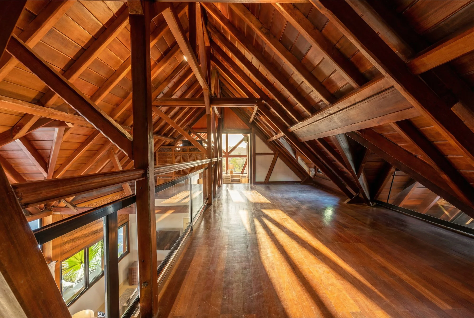 Intérieur d'une maison en bois avec un plafond en pente soutenu par des poutres en bois, lumière naturelle entrant par de grandes fenêtres, sol en bois, ambiance chaleureuse et spacieuse.