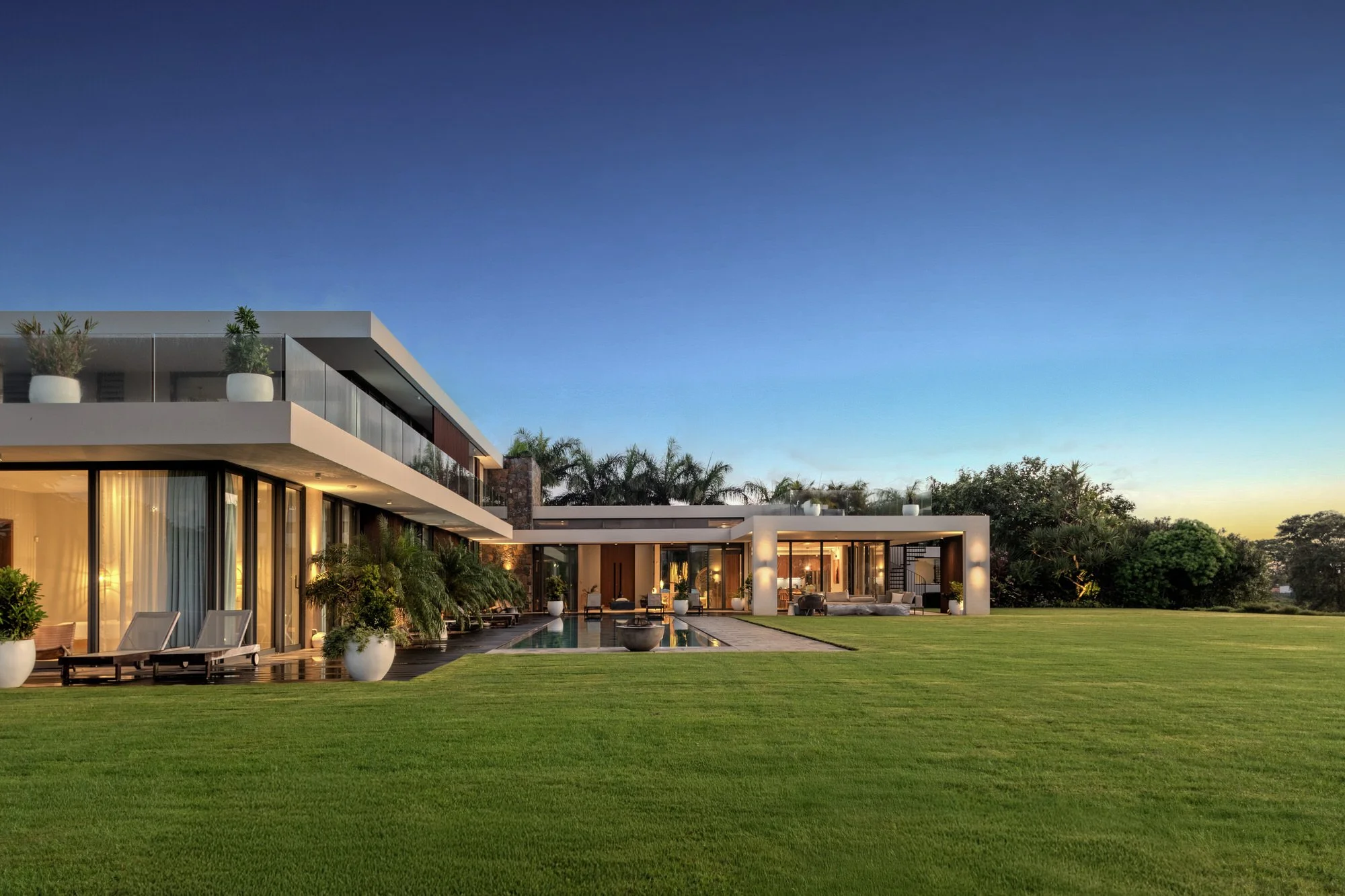 Villa moderne avec piscine, jardin bien entretenu, grande baie vitrée, mobilier extérieur et décorations végétales, sous un ciel bleu en soirée.